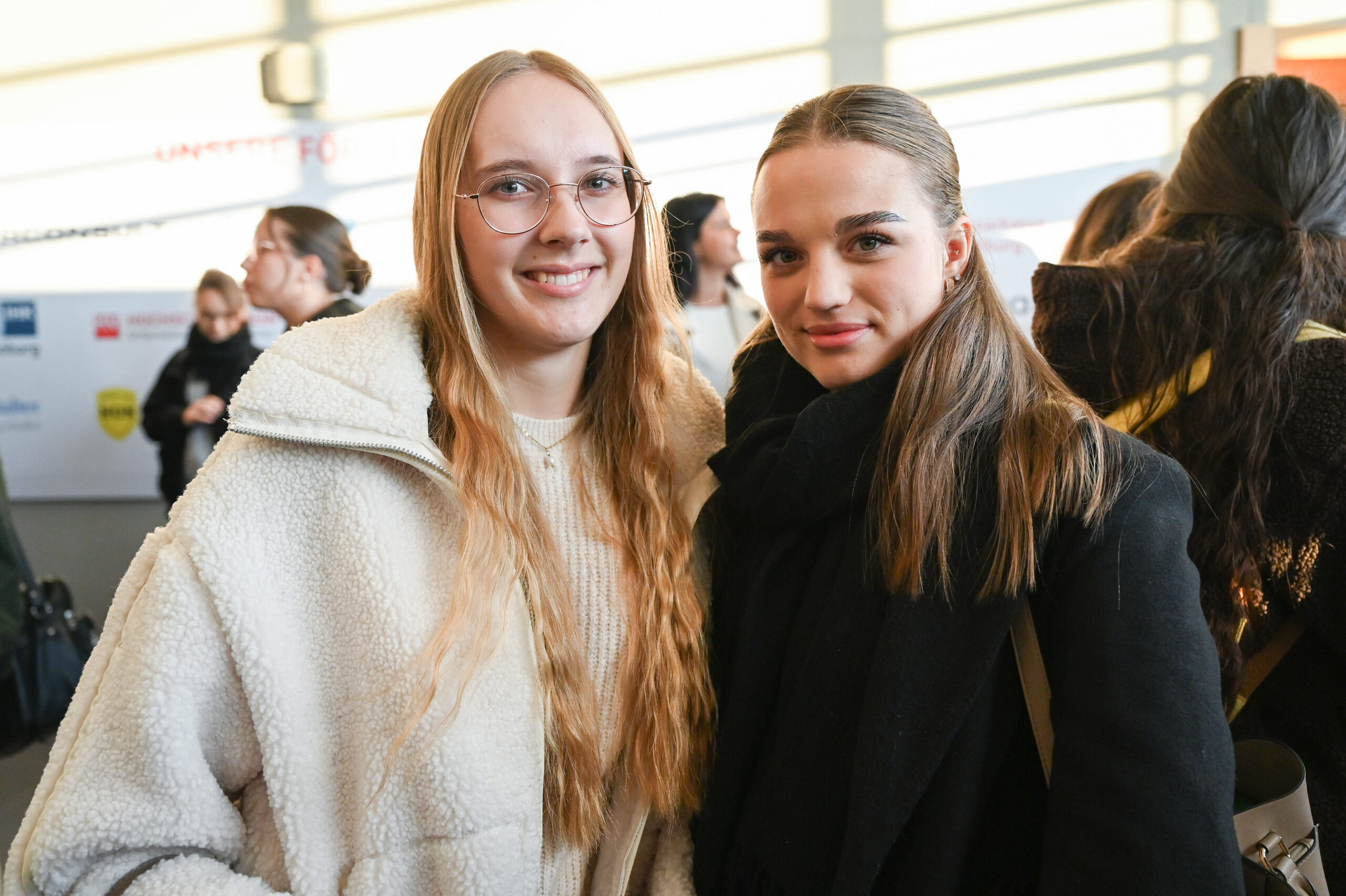 Zwei junge Frauen posieren zusammen und lächeln bei einer Indoor-Veranstaltung. Die eine, in einem cremefarbenen Mantel, hat langes blondes Haar und eine Brille; die andere trägt einen schwarzen Mantel und ihr dunkelblondes Haar zurückgebunden. Es ist ein automatisch gespeicherter Moment - ein automatisch gespeicherter Entwurf. at Hochschule Coburg