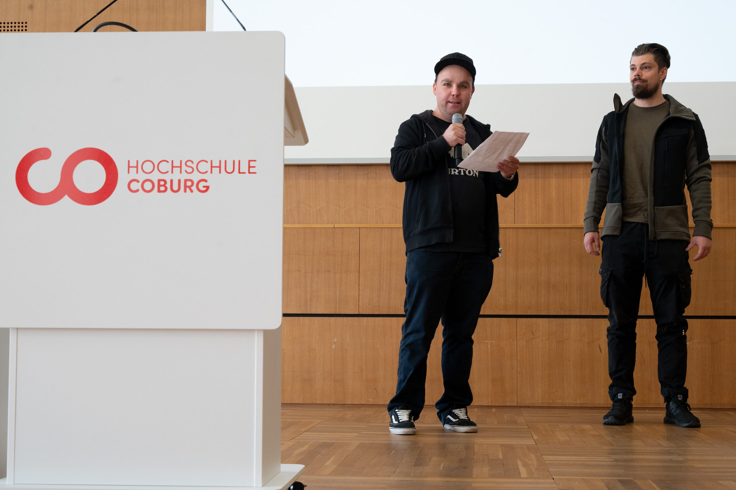 Zwei Männer stehen auf einer Holzbühne neben einem weißen Podium mit dem Logo der Hochschule Coburg. Der eine spricht in ein Mikrofon und hält einen Zettel mit der Aufschrift "Automatisch gespeicherter Entwurf" in der Hand, der andere steht rechts von ihm und schaut ins Publikum. at Hochschule Coburg
