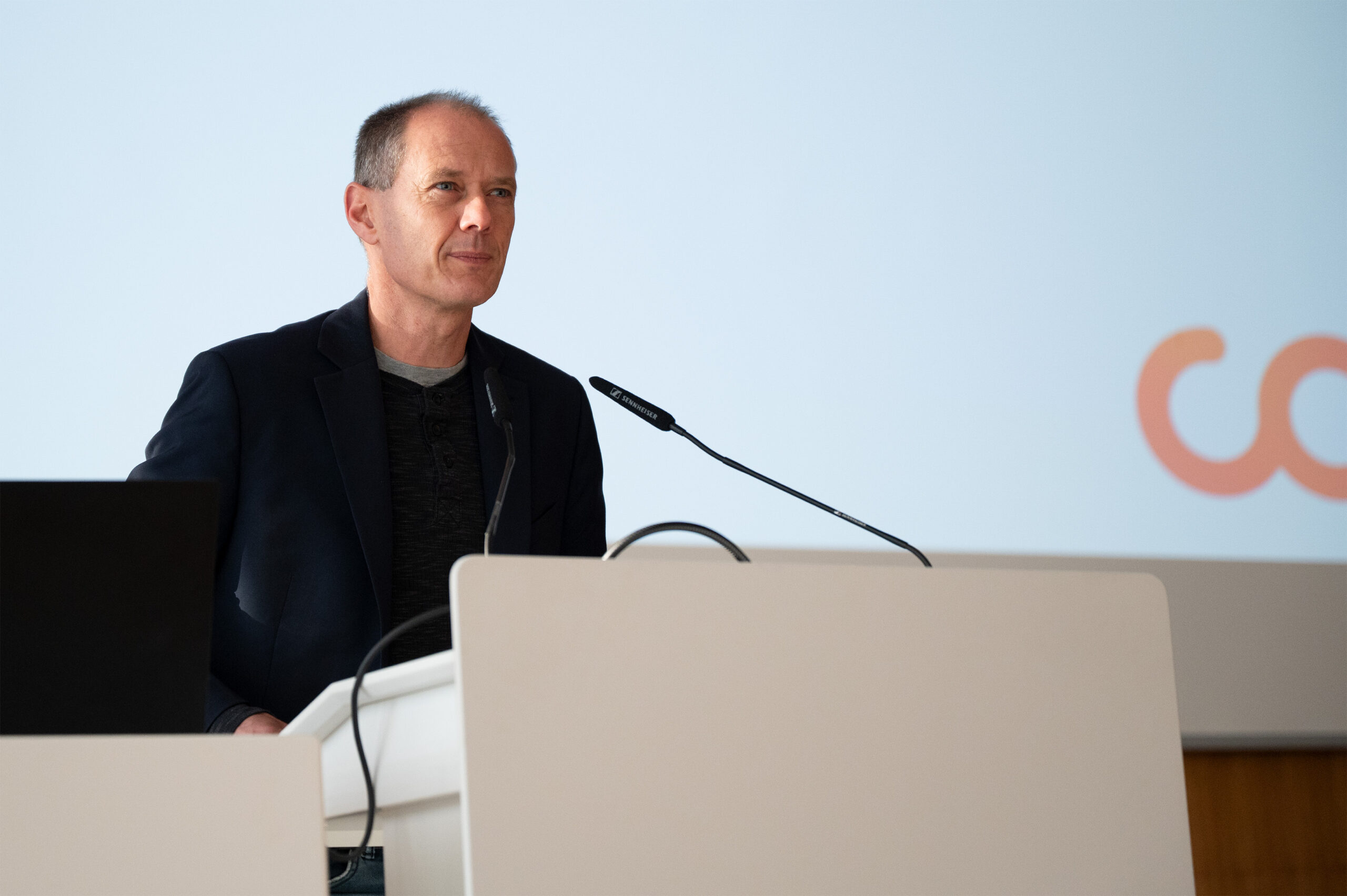 Ein Mann in einem dunklen Blazer steht an einem weißen Podium und spricht in einem hellen Raum in ein Mikrofon. Auf einem Laptop wird ein automatisch gespeicherter Entwurf angezeigt, während auf der Projektionsfläche hinter ihm Mikrofone und ein Teil eines Logos zu sehen sind. at Hochschule Coburg