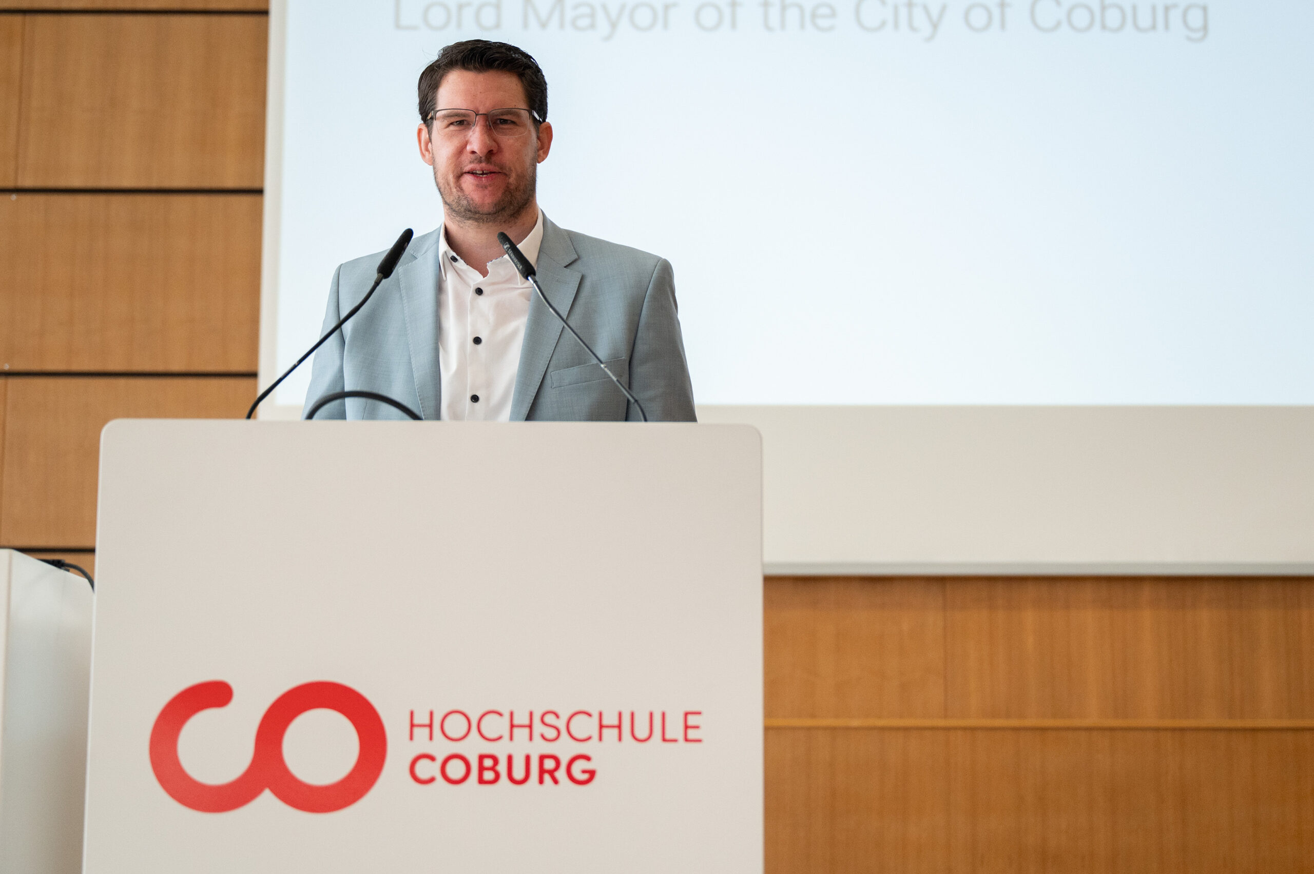 Ein Mann in einem hellgrauen Anzug steht an einem Podium mit dem Logo der Hochschule Coburg und spricht vor einem Bildschirm mit teilweise sichtbarem Text in Englisch und Deutsch. Im Hintergrund ist eine Holzvertäfelung zu sehen. at Hochschule Coburg