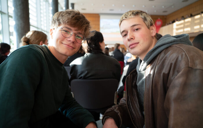 Zwei junge Männer sitzen eng beieinander in einem sonnenbeschienenen Raum und lächeln in die Kamera. Andere Personen sitzen in Reihen hinter ihnen, was auf eine Gruppenveranstaltung oder ein Treffen hindeutet. Die Atmosphäre wirkt entspannt und freundlich. at Hochschule Coburg