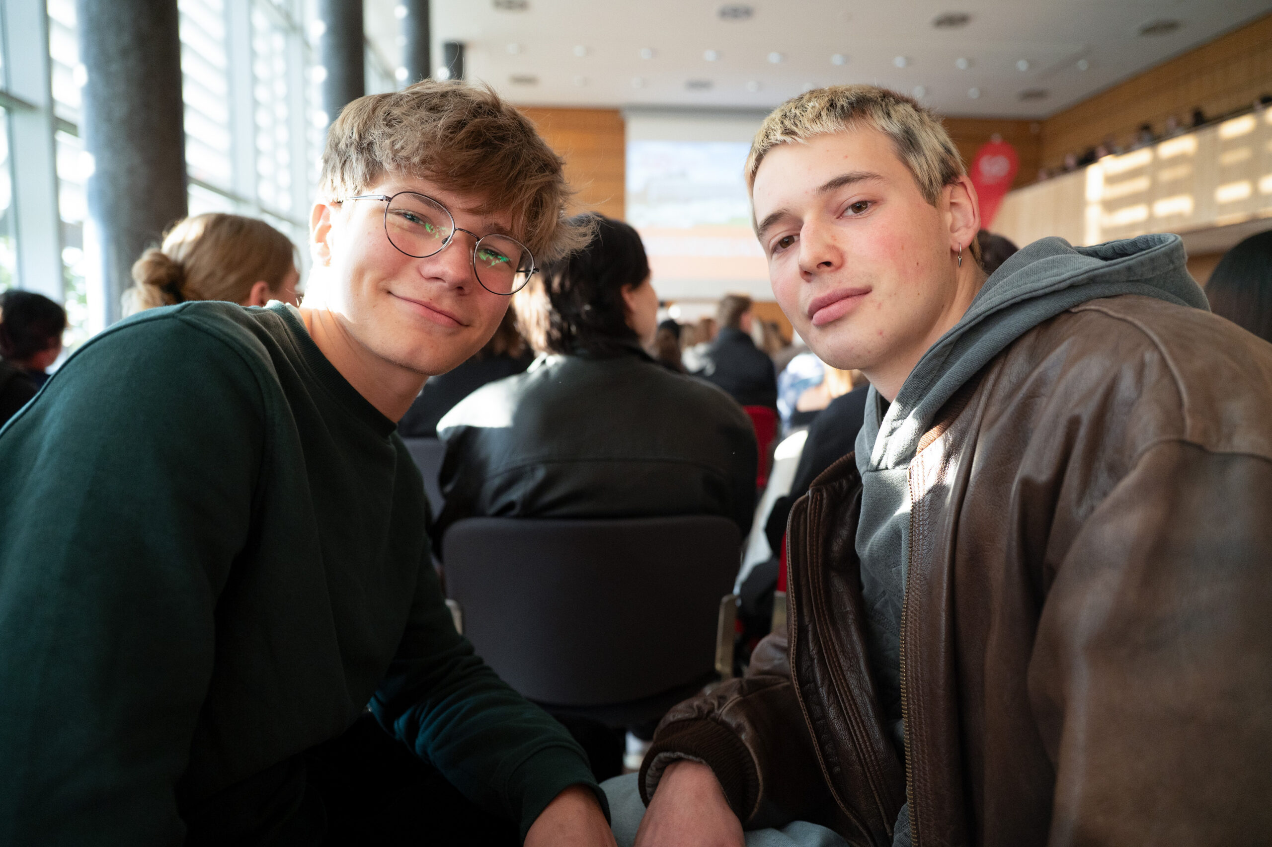 Zwei junge Männer sitzen eng beieinander in einem sonnenbeschienenen Raum und lächeln in die Kamera. Andere Personen sitzen in Reihen hinter ihnen, was auf eine Gruppenveranstaltung oder ein Treffen hindeutet. Die Atmosphäre wirkt entspannt und freundlich. at Hochschule Coburg