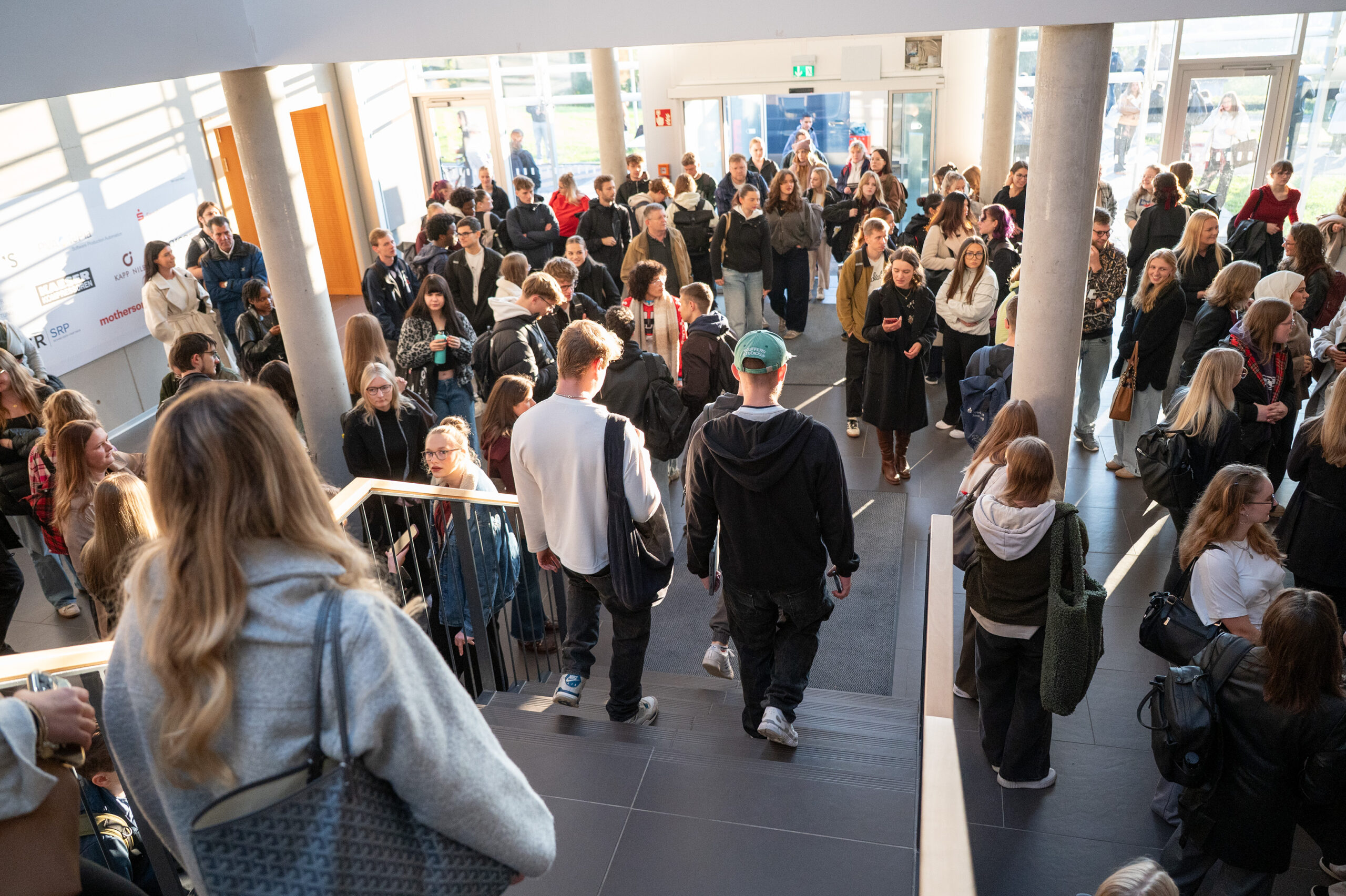 Eine große Gruppe von Menschen, meist junge Erwachsene, versammelt sich in einem hellen, modernen Gebäude. Einige gehen die Treppe hinunter, während andere in der belebten Lobby stehen und sich unterhalten, ihre Gespräche wie ein automatisch gespeicherter Entwurf - frisch und voller Potenzial. at Hochschule Coburg