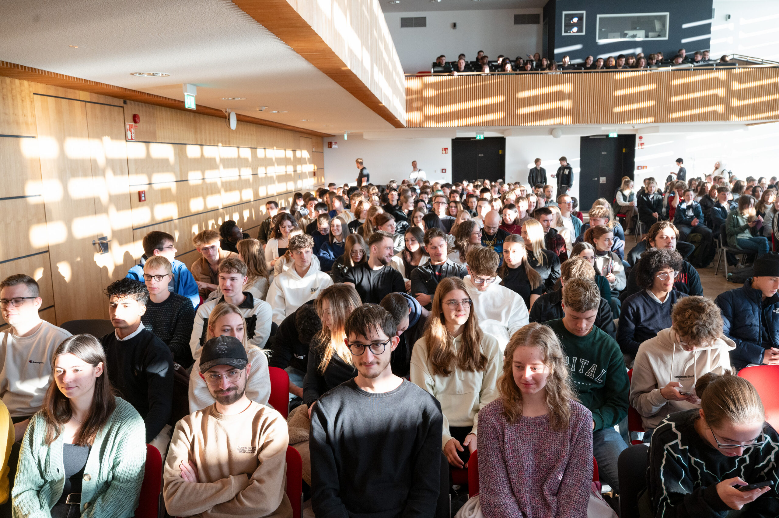 Eine große Gruppe von Studenten sitzt in einem hellen, sonnendurchfluteten Hörsaal dicht an dicht in Stuhlreihen. Einige blicken nach vorne, andere unterhalten sich. Das Sonnenlicht wirft gitterartige Schatten auf die Wand und die Menschen, wie ein automatisch gespeicherter Entwurf ihrer Zusammenkunft. at Hochschule Coburg