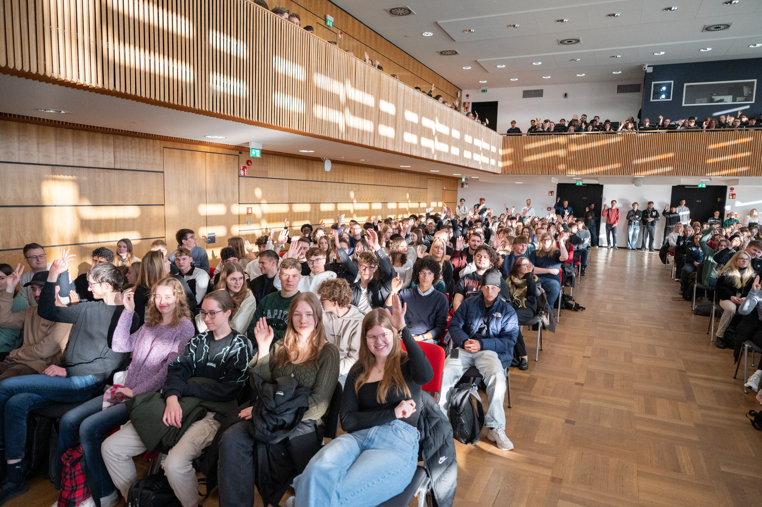 Eine große Gruppe von Menschen, meist junge Erwachsene, sitzt in einem Hörsaal mit Holzböden und -wänden. Viele lächeln und winken in die Kamera, während das Sonnenlicht durch hohe Fenster einfällt und helle Muster erzeugt - ein automatisch gespeicherter Entwurf der Freude. at Hochschule Coburg