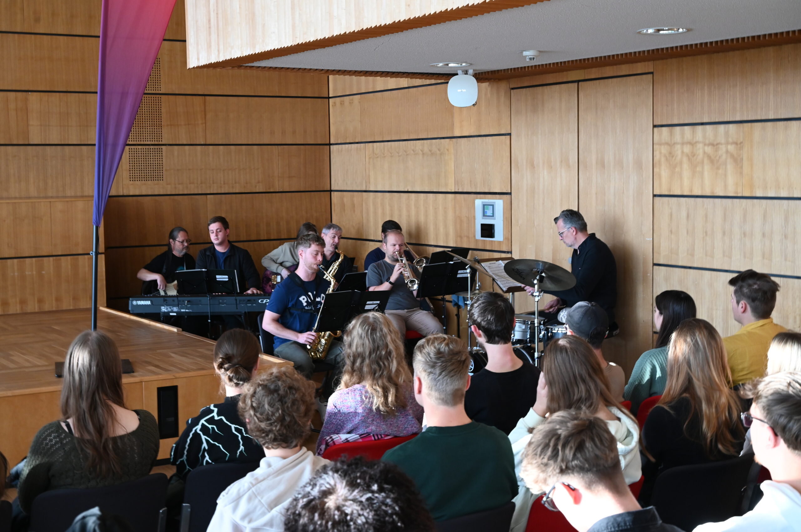 Eine Jazzband mit sechs Musikern, darunter ein Pianist, ein Schlagzeuger, ein Saxophonist und ein Posaunist, spielt auf einer Holzbühne vor einem aufmerksamen sitzenden Publikum in einem modernen Raum. at Hochschule Coburg