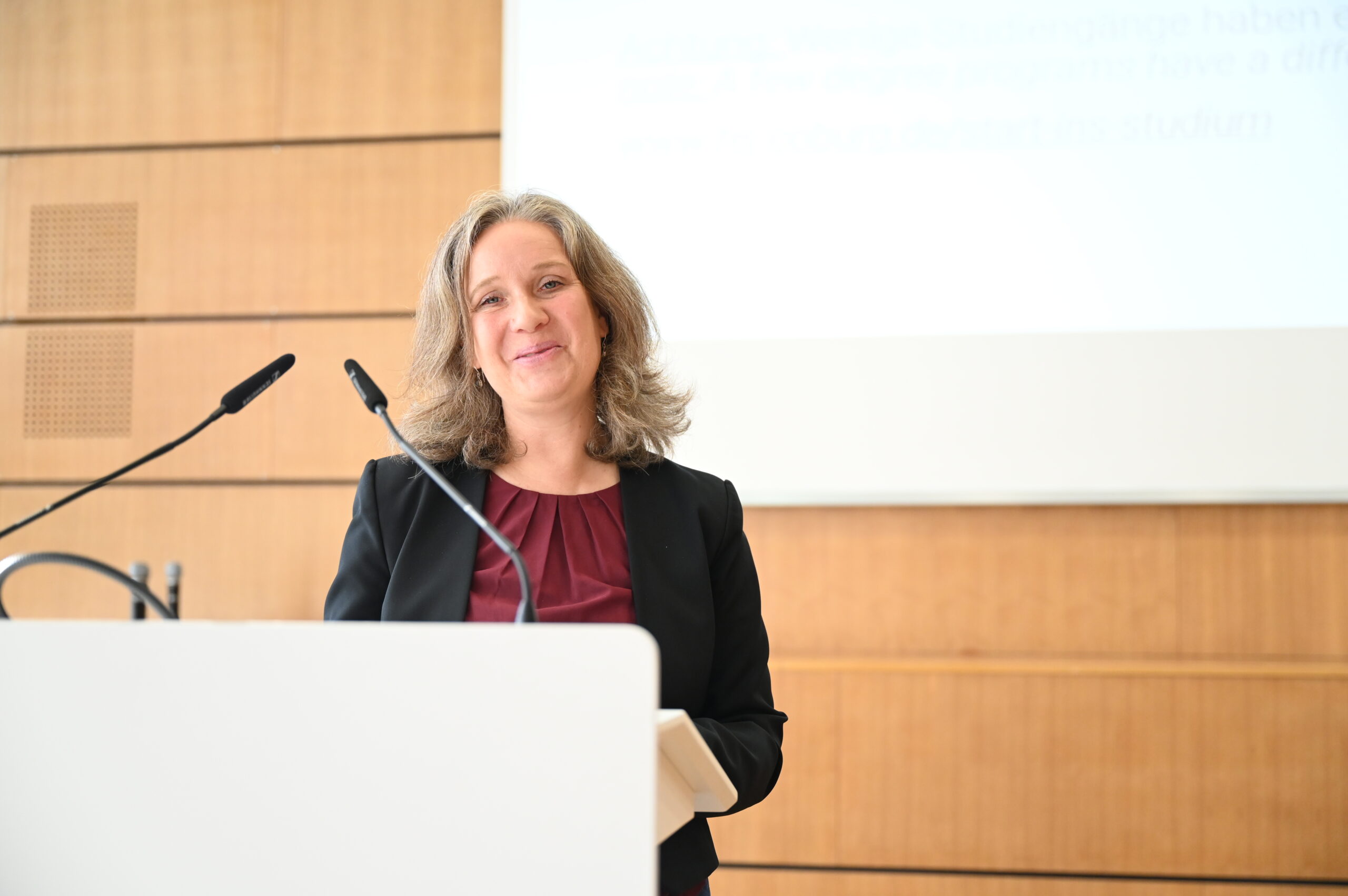 Eine Frau mit schulterlangem blondem Haar, schwarzem Blazer und weinrotem Oberteil steht an einem Podium mit Mikrofonen in einem gut beleuchteten, holzgetäfelten Raum, hinter dem sich ein Bildschirm befindet. at Hochschule Coburg