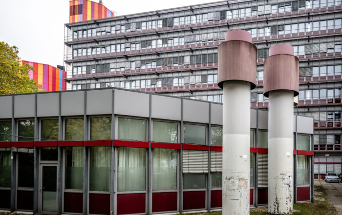 Ein modernes Gebäude mit großen Fenstern und roten Akzenten steht vor einem höheren Bau, in dem innovative Konzepte und Lernräume der Zukunft für Studierende präsentiert werden. In der Nähe befinden sich zwei große zylindrische Lüftungsschächte, links davon stehen Bäume. at Hochschule Coburg