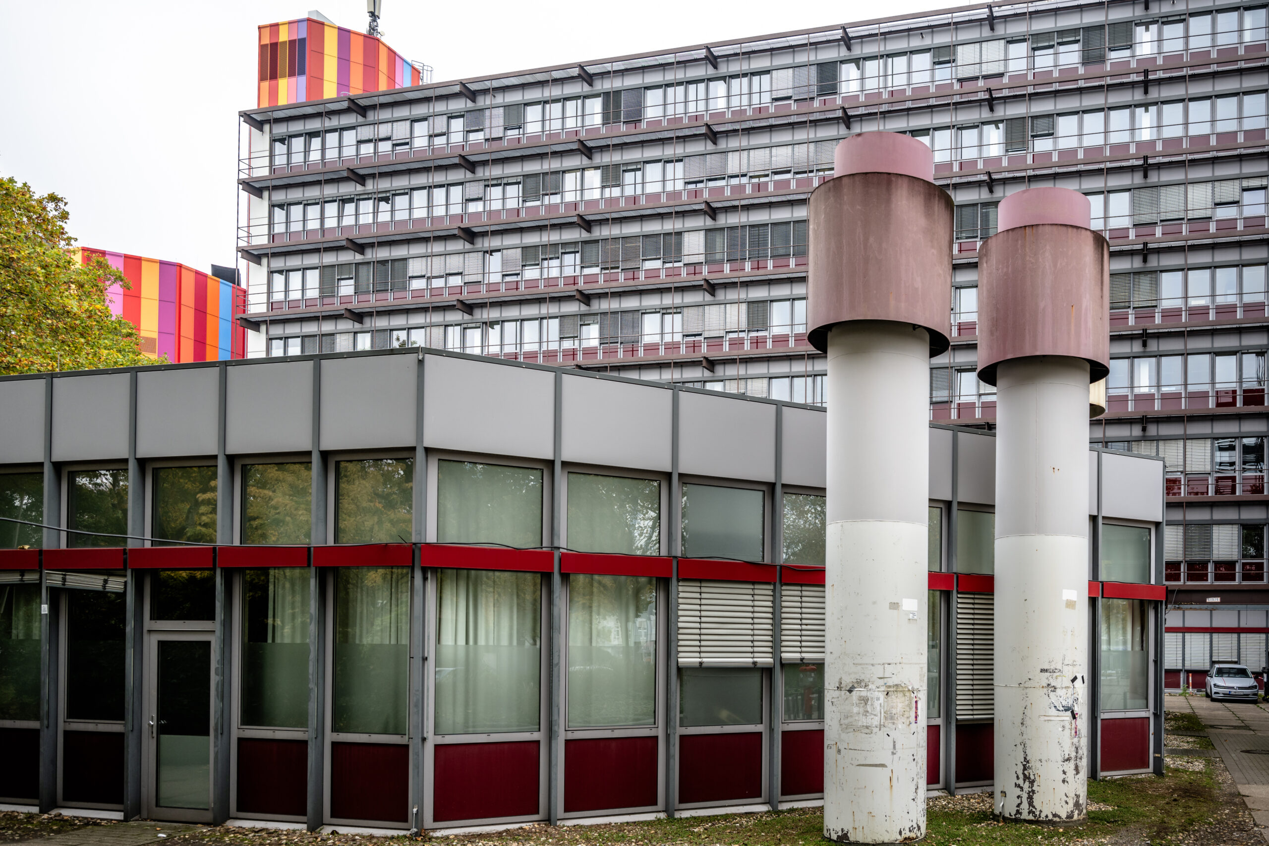 Ein modernes Gebäude mit großen Fenstern und roten Akzenten steht vor einem höheren Bau, in dem innovative Konzepte und Lernräume der Zukunft für Studierende präsentiert werden. In der Nähe befinden sich zwei große zylindrische Lüftungsschächte, links davon stehen Bäume. at Hochschule Coburg