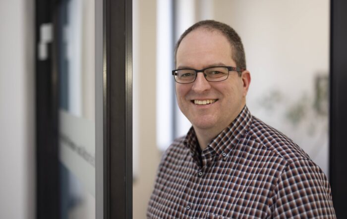 Ein Mann mit kurzen Haaren und Brille, der ein kariertes Hemd trägt, steht lächelnd neben einer Glastür in einem hellen Innenraum - vielleicht der Experimentalphysiker Uphues nach der Verleihung eines Wirtschaftspreises. at Hochschule Coburg