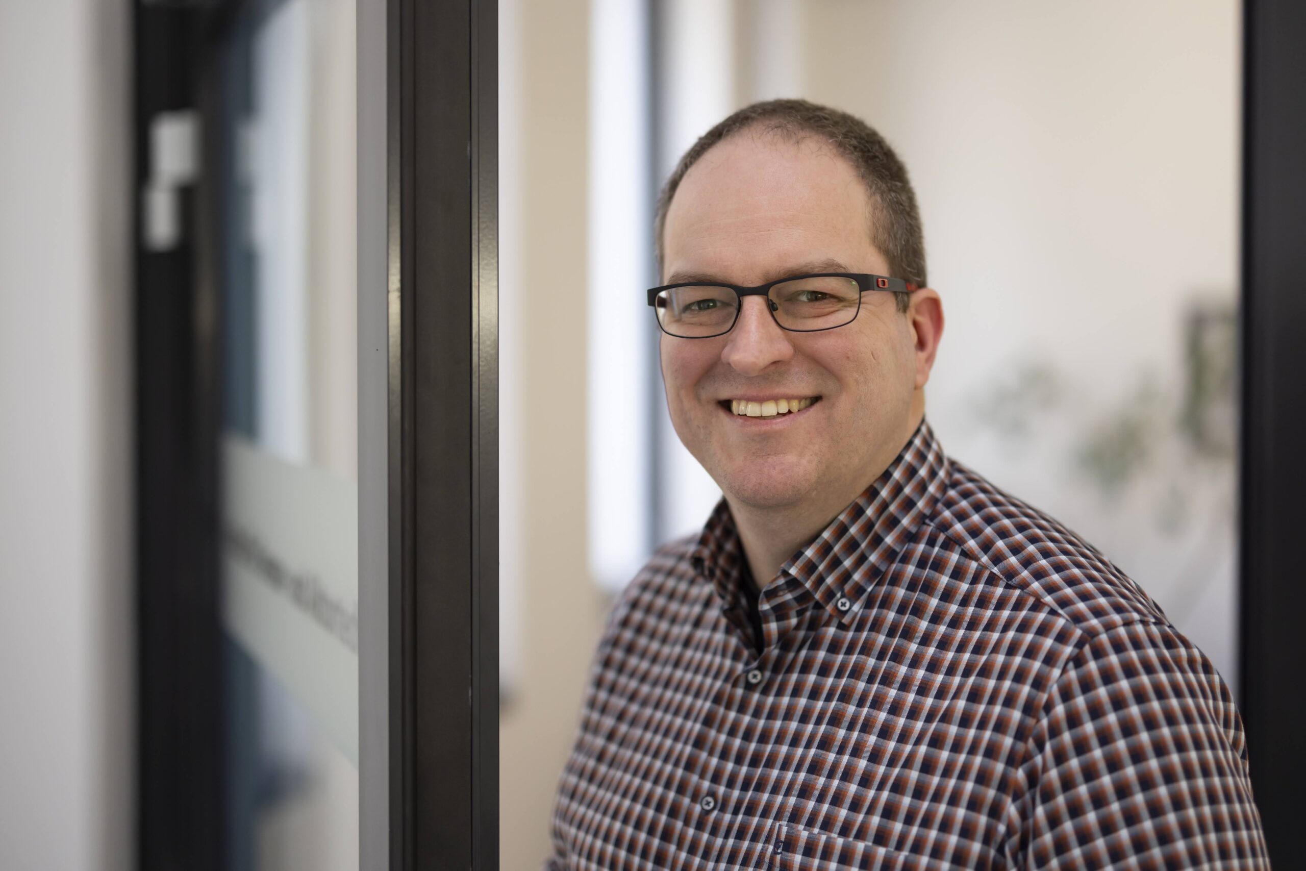 Ein Mann mit kurzen Haaren und Brille, der ein kariertes Hemd trägt, steht lächelnd neben einer Glastür in einem hellen Innenraum - vielleicht der Experimentalphysiker Uphues nach der Verleihung eines Wirtschaftspreises. at Hochschule Coburg