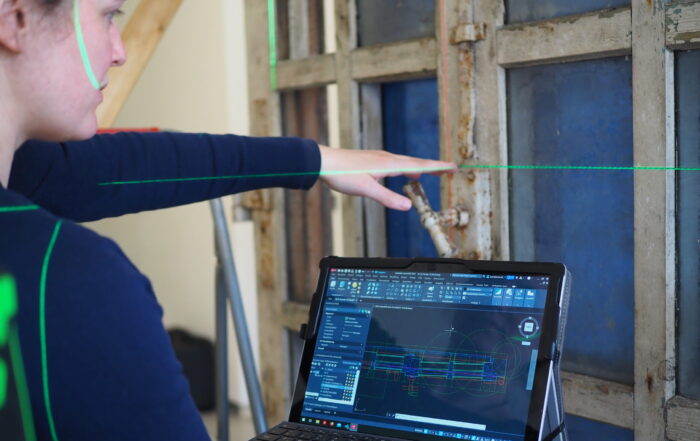 Eine Person benutzt einen Laptop, auf dem technische CAD-Zeichnungen und ein automatisch gespeicherter Entwurf angezeigt werden, während sie auf einen alten Fensterrahmen zeigt. Es sind grüne Laserlinien zu sehen, die bei präzisen Messungen oder Ausrichtungen behilflich sind. at Hochschule Coburg