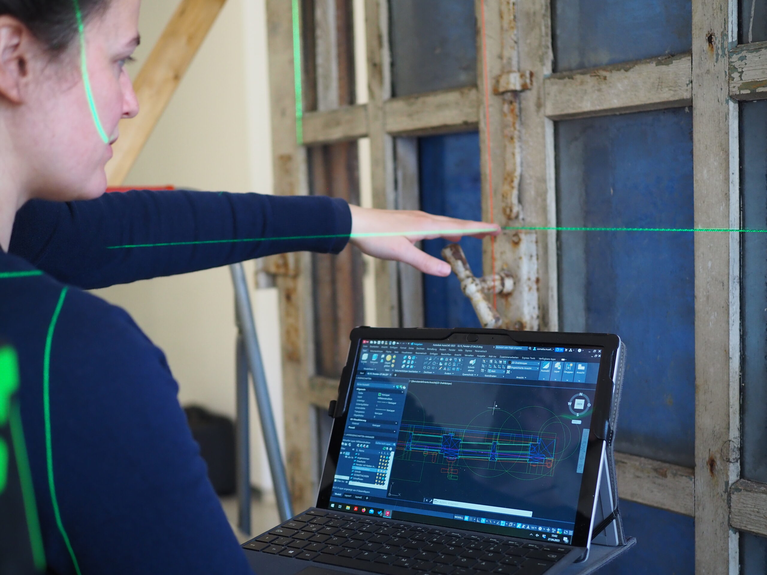 Eine Person benutzt einen Laptop, auf dem technische CAD-Zeichnungen und ein automatisch gespeicherter Entwurf angezeigt werden, während sie auf einen alten Fensterrahmen zeigt. Es sind grüne Laserlinien zu sehen, die bei präzisen Messungen oder Ausrichtungen behilflich sind. at Hochschule Coburg