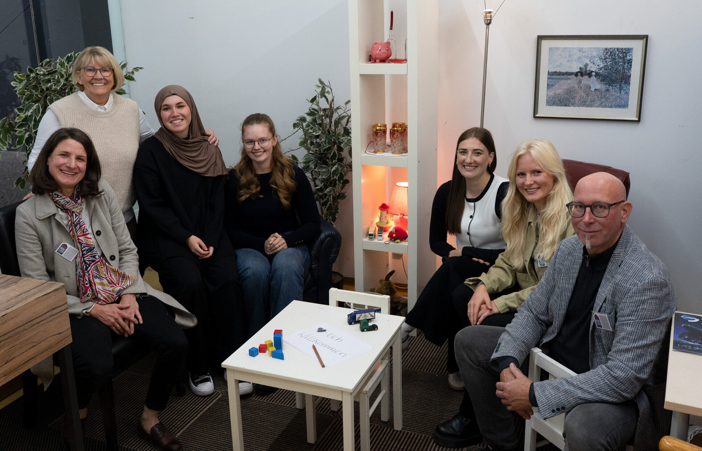 Eine Gruppe von sieben Personen, darunter Studis, vier Frauen sitzend, ein Mann sitzend und zwei Frauen stehend, posieren und lächeln in einem gemütlichen, gut beleuchteten Raum mit Pflanzen, Regalen und einem kleinen Tisch mit bunten Blöcken in der Mitte. Die Atmosphäre ist echt herzlich. at Hochschule Coburg