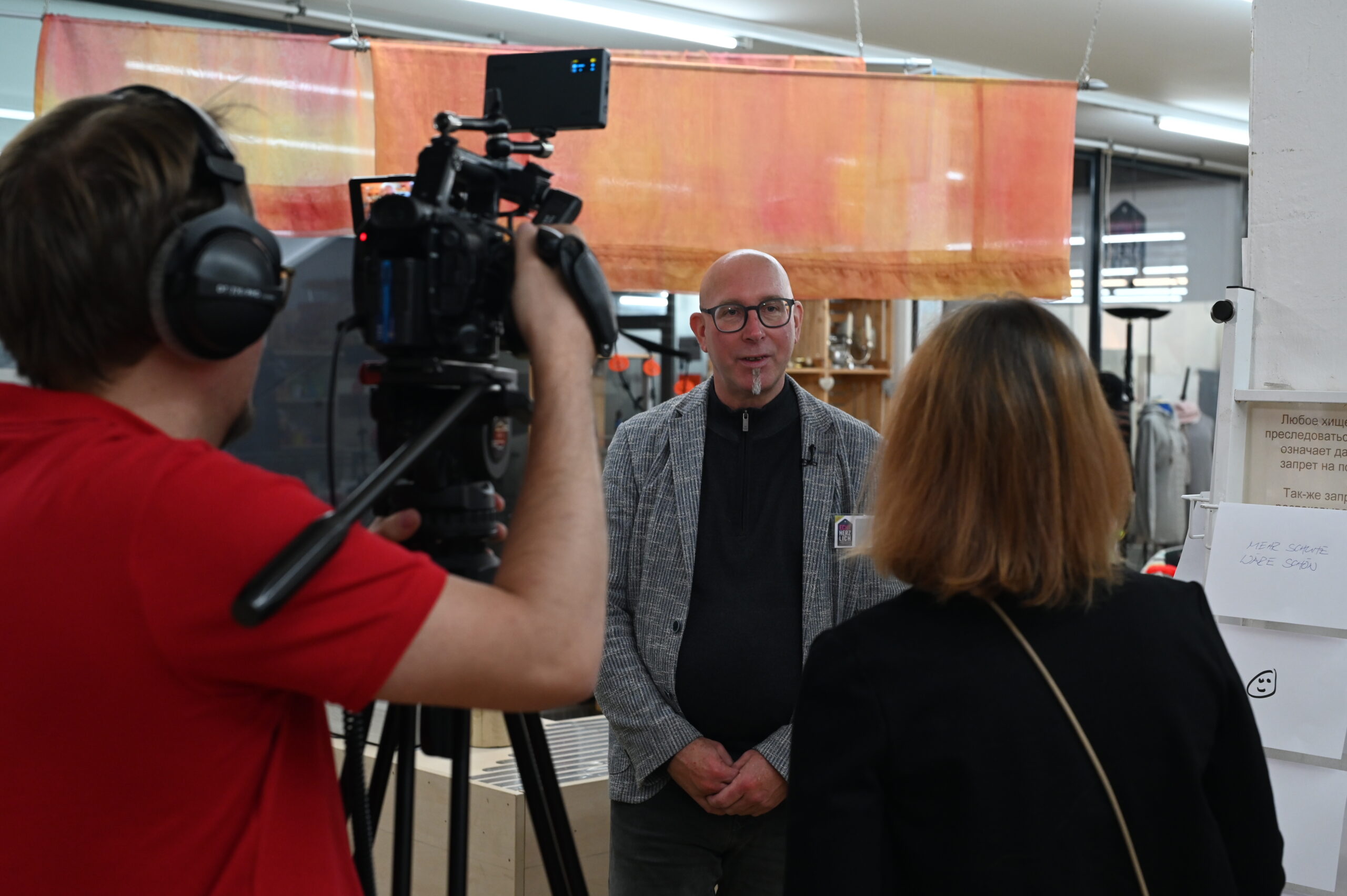 Ein Mann steht vor einer Kamera in einem Innenraum und spricht mit einer Frau, während eine andere Person mit einer professionellen Videoausrüstung aufnimmt. Zu sehen sind Regale und Stoffbehänge, die ein echt herzliches Einkaufserlebnis im Hintergrund schaffen. at Hochschule Coburg