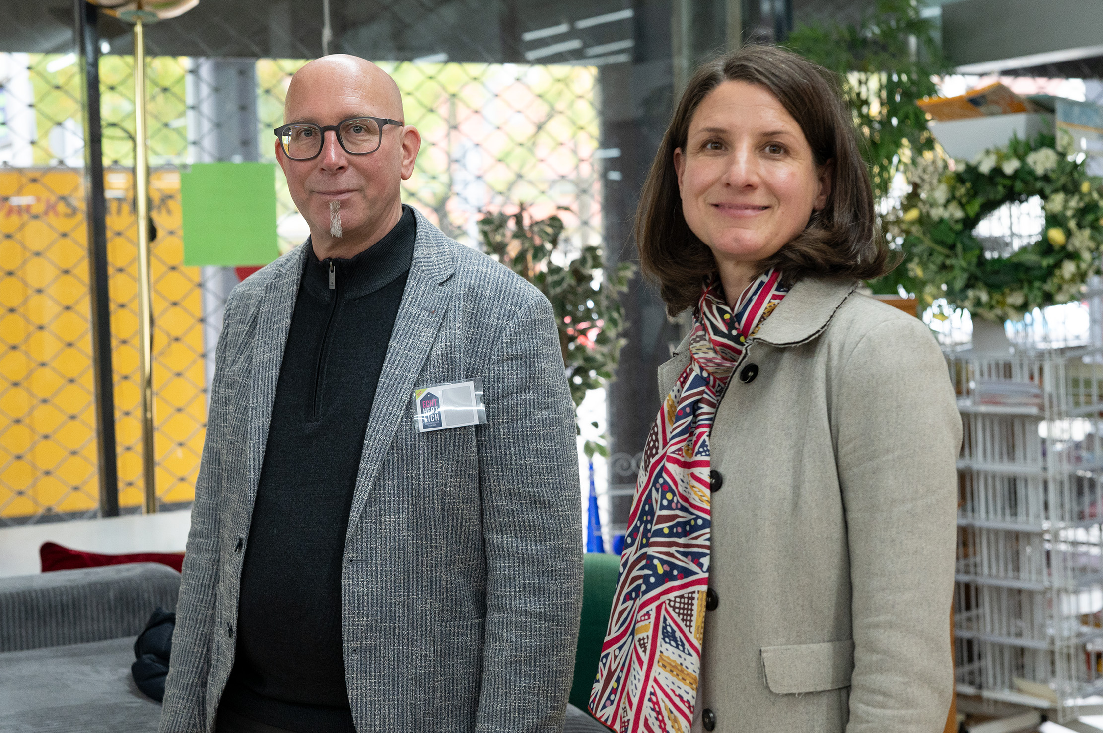 Ein Mann in einem grauen Blazer und mit Brille steht neben einer Frau in einem hellen Mantel mit einem bunten Schal. Sie sind drinnen, lächelnd, mit Pflanzen und Regalen in der Nähe - ein echt herzliches Einkaufserlebnis für Studis. at Hochschule Coburg