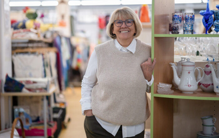 Eine ältere Frau mit Brille und beigem Pullover lächelt und macht ein echt herzliches Einkaufserlebnis in einem Laden, in dem bunte Gläser, Teekannen und andere Schätze in den Regalen stehen - ein einladender Ort für Studis und Einheimische gleichermaßen. at Hochschule Coburg
