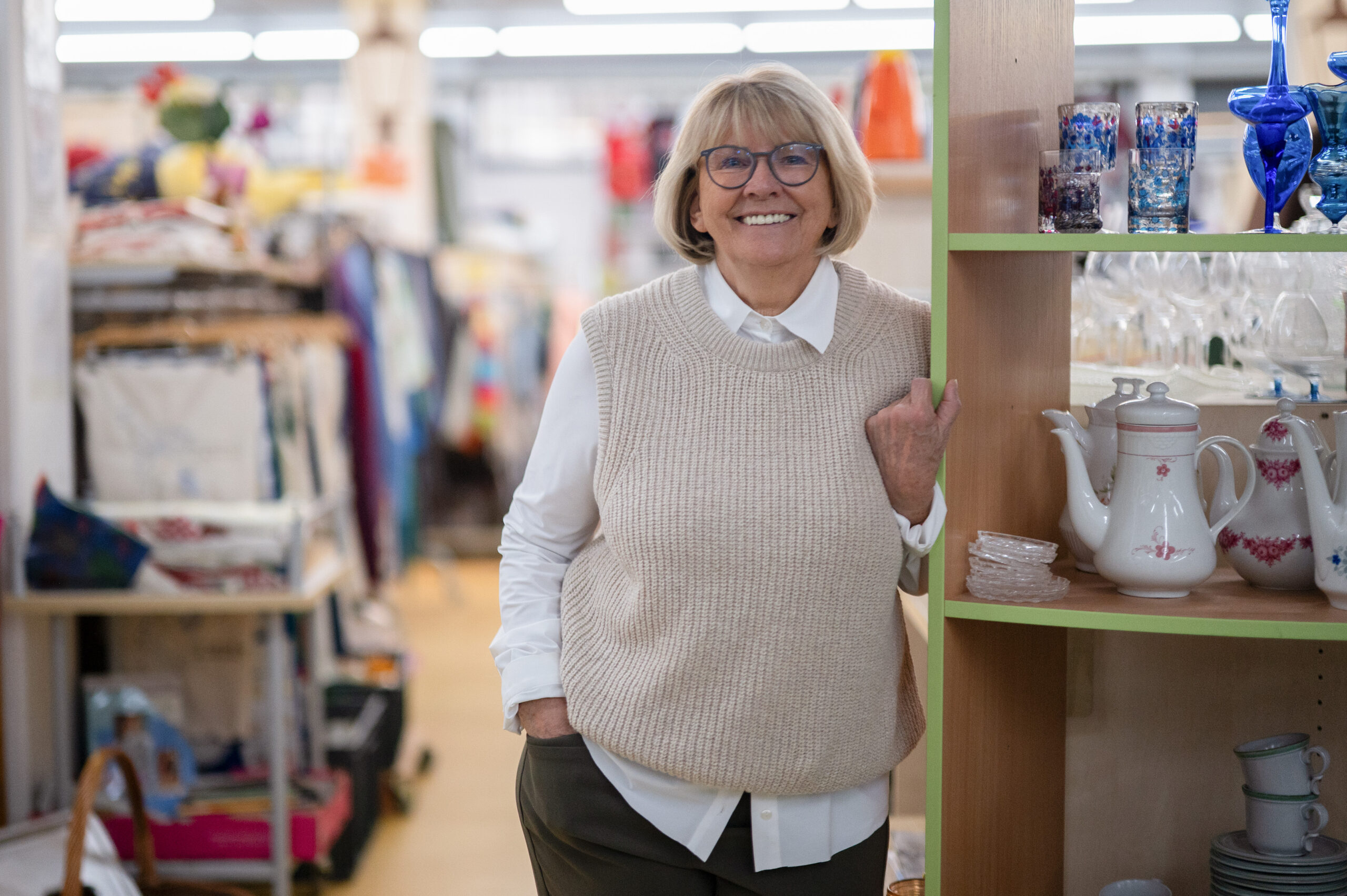 Eine ältere Frau mit Brille und beigem Pullover lächelt und macht ein echt herzliches Einkaufserlebnis in einem Laden, in dem bunte Gläser, Teekannen und andere Schätze in den Regalen stehen - ein einladender Ort für Studis und Einheimische gleichermaßen. at Hochschule Coburg