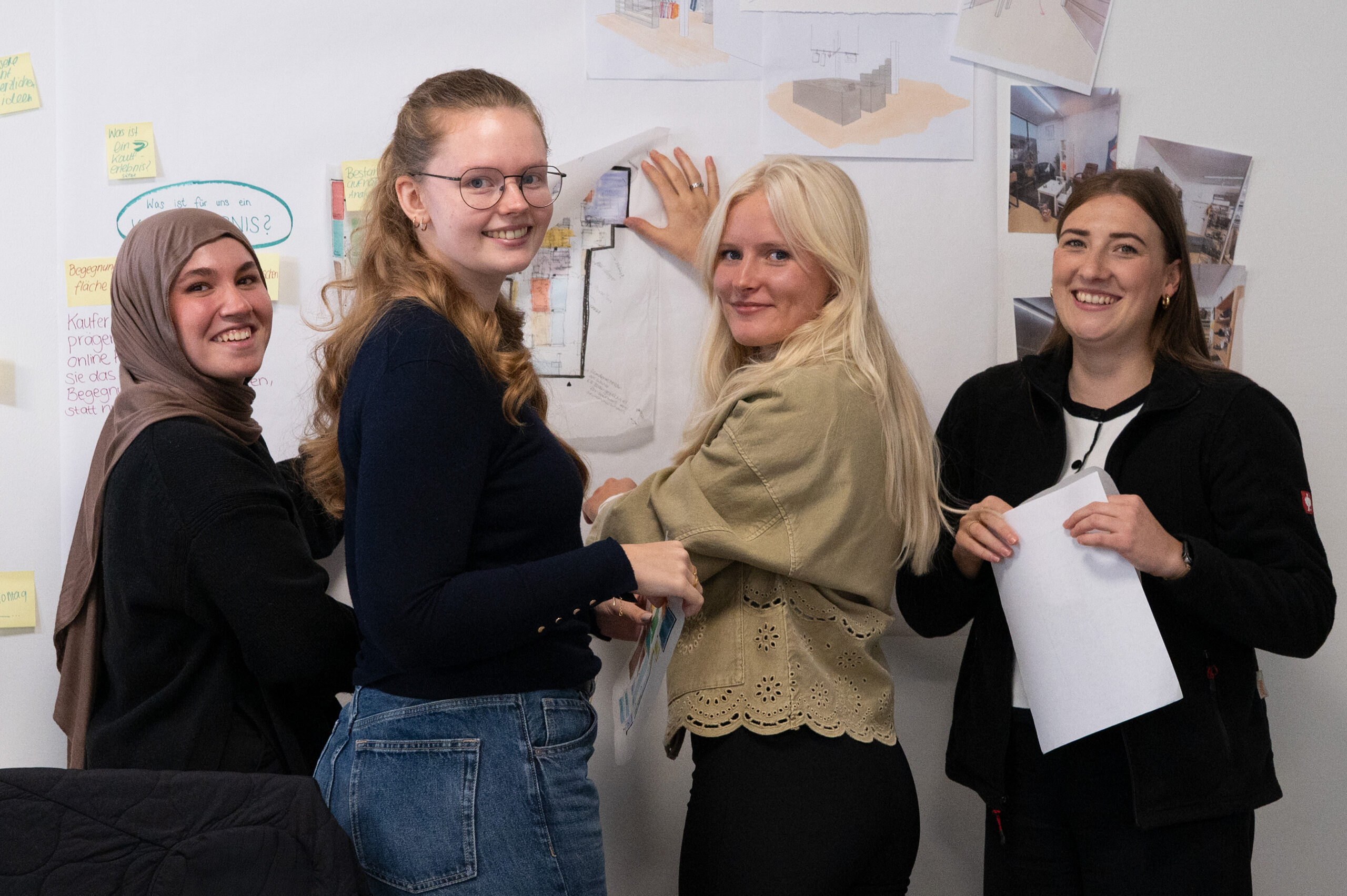 Vier Frauen, die sehr herzlich und kooperativ wirken, stehen lächelnd vor einer Wand, die mit Skizzen, Notizen und Diagrammen bedeckt ist. Eine Frau zeigt auf eine Zeichnung, während die anderen in die Kamera schauen, die engagierte Studis bei der Arbeit zeigt. at Hochschule Coburg