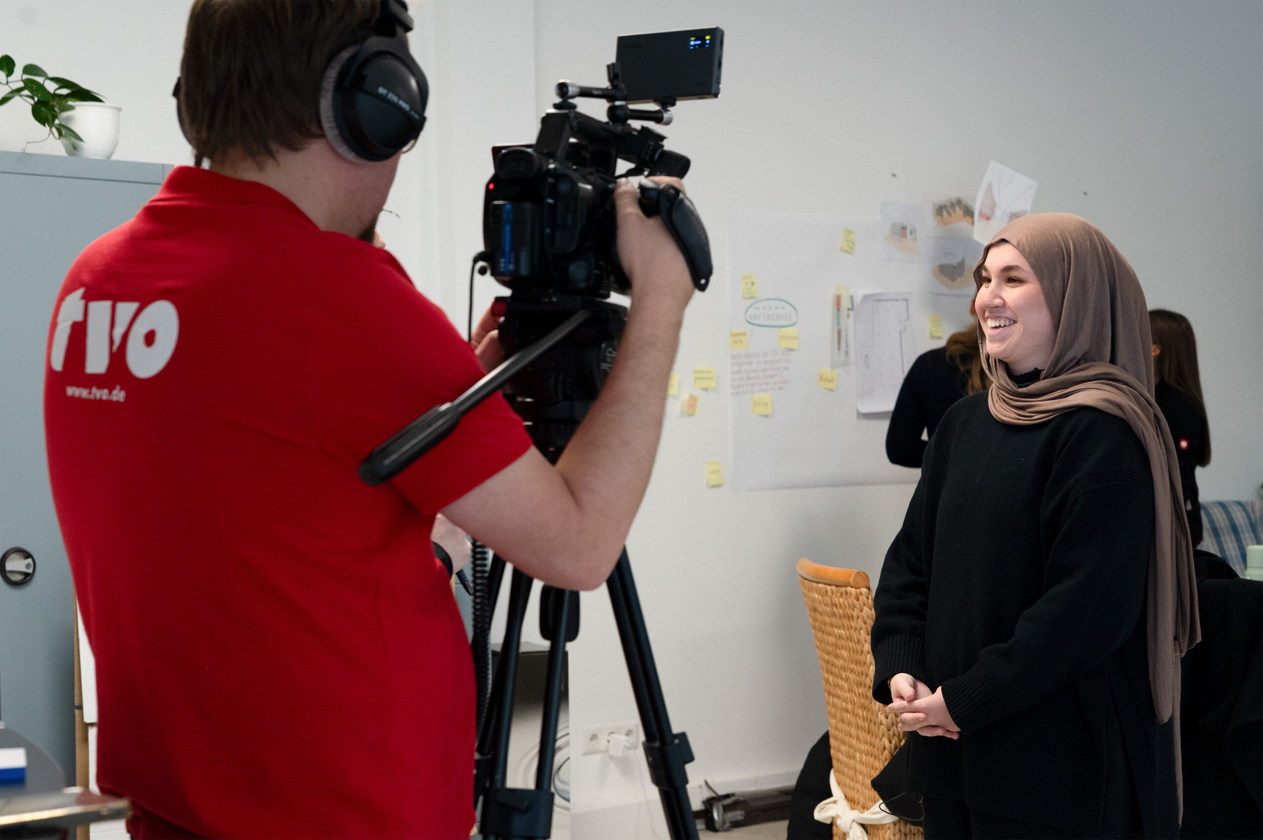 Eine Person in einem roten Hemd filmt eine lächelnde Frau im Hidschab mit einer Videokamera in einer Studiokulisse, während an der Wand im Hintergrund Notizen und Skizzen zu sehen sind. at Hochschule Coburg
