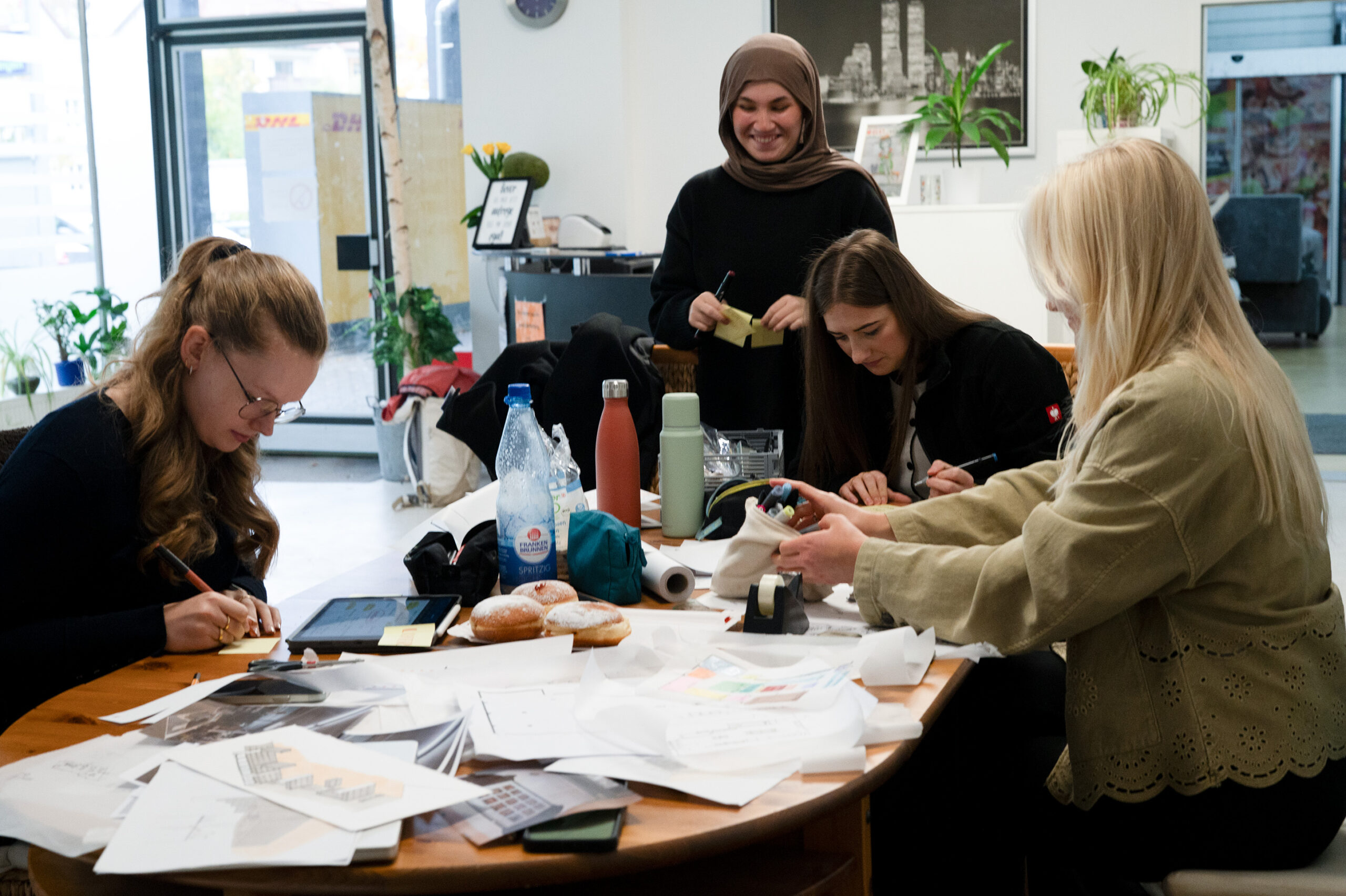 Vier Studis sitzen um einen mit Papieren, Heften und Getränken bedeckten Tisch und arbeiten und schreiben. Eine Frau steht lächelnd im Hintergrund. Die Atmosphäre ist echt herzlich, gemeinschaftlich und kreativ in einem hellen Innenraum. at Hochschule Coburg