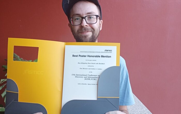 Ein Mann mit Brille und Mütze lächelt, während er eine Urkundenmappe mit dem Preis für das beste Poster für Forschung über faire Suchmaschinen in der Hand hält und neben einer roten Wand und Pflanzen steht. at Hochschule Coburg