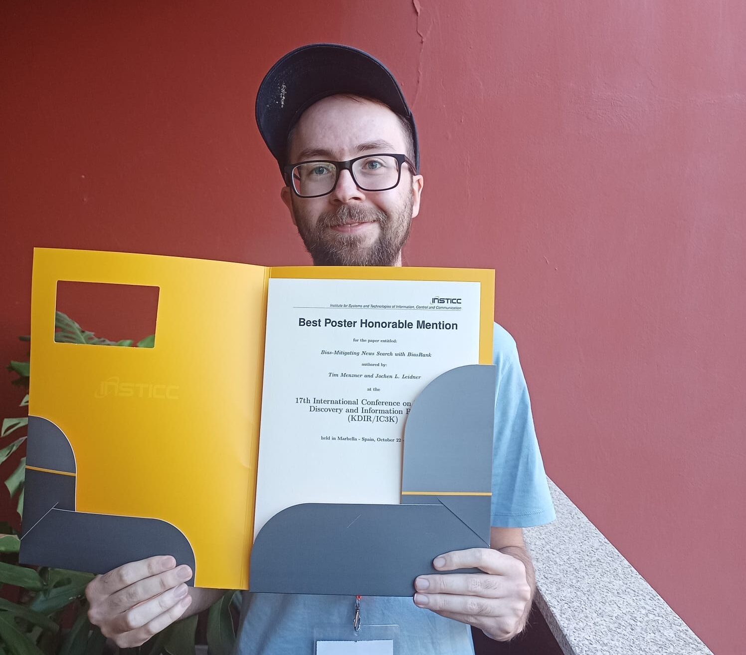 Ein Mann mit Brille und Mütze lächelt, während er eine Urkundenmappe mit dem Preis für das beste Poster für Forschung über faire Suchmaschinen in der Hand hält und neben einer roten Wand und Pflanzen steht. at Hochschule Coburg