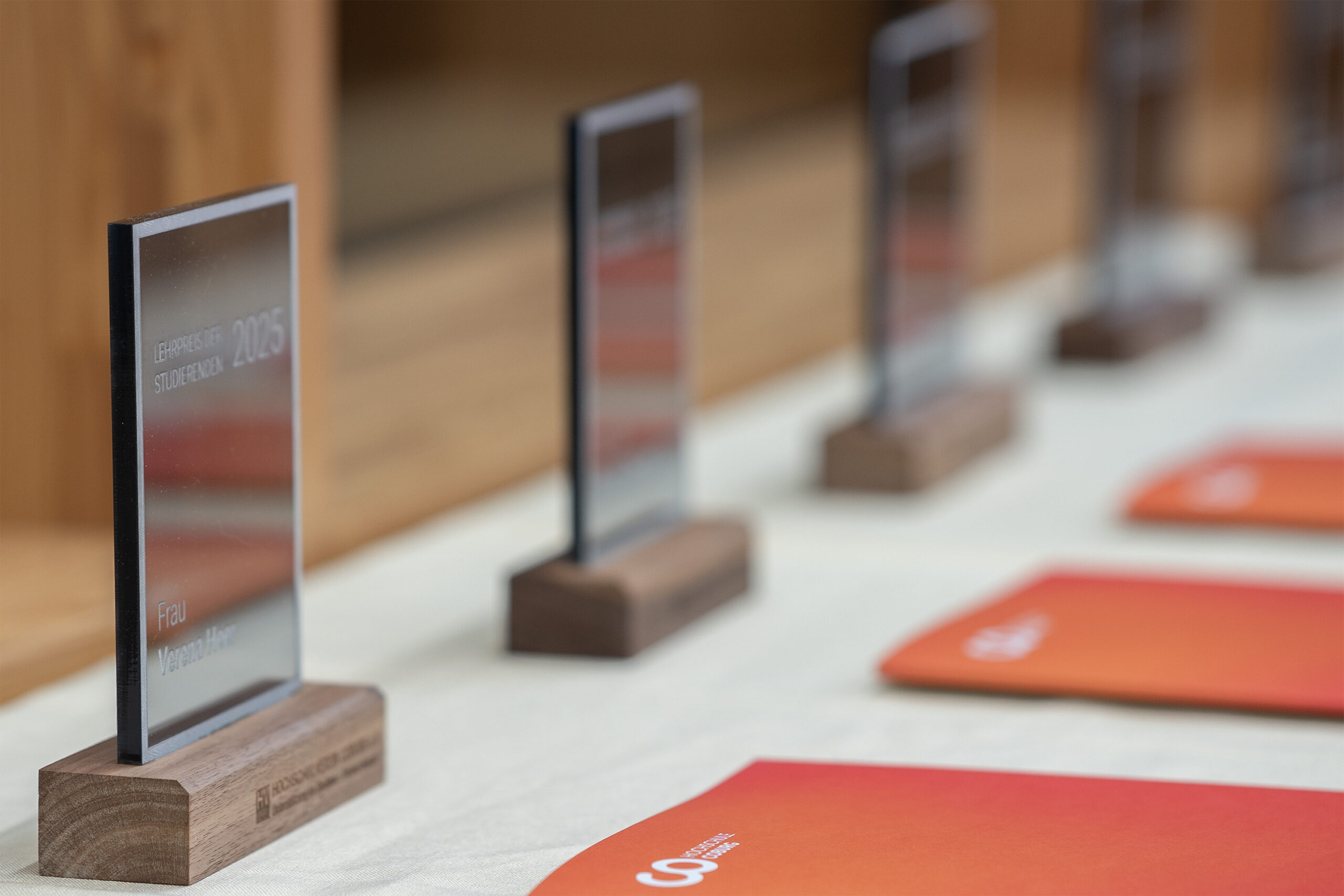 Mehrere gläserne Auszeichnungen mit hölzernen Sockeln sind auf einem Tisch aufgereiht, neben einer Reihe von orangefarbenen Mappen, bereit, um bei der Zeremonie beliebteste Lehrende und innovative Lehre zu ehren. Der Text auf den Auszeichnungen ist teilweise sichtbar, und die Szene findet in einem Innenraum statt. at Hochschule Coburg