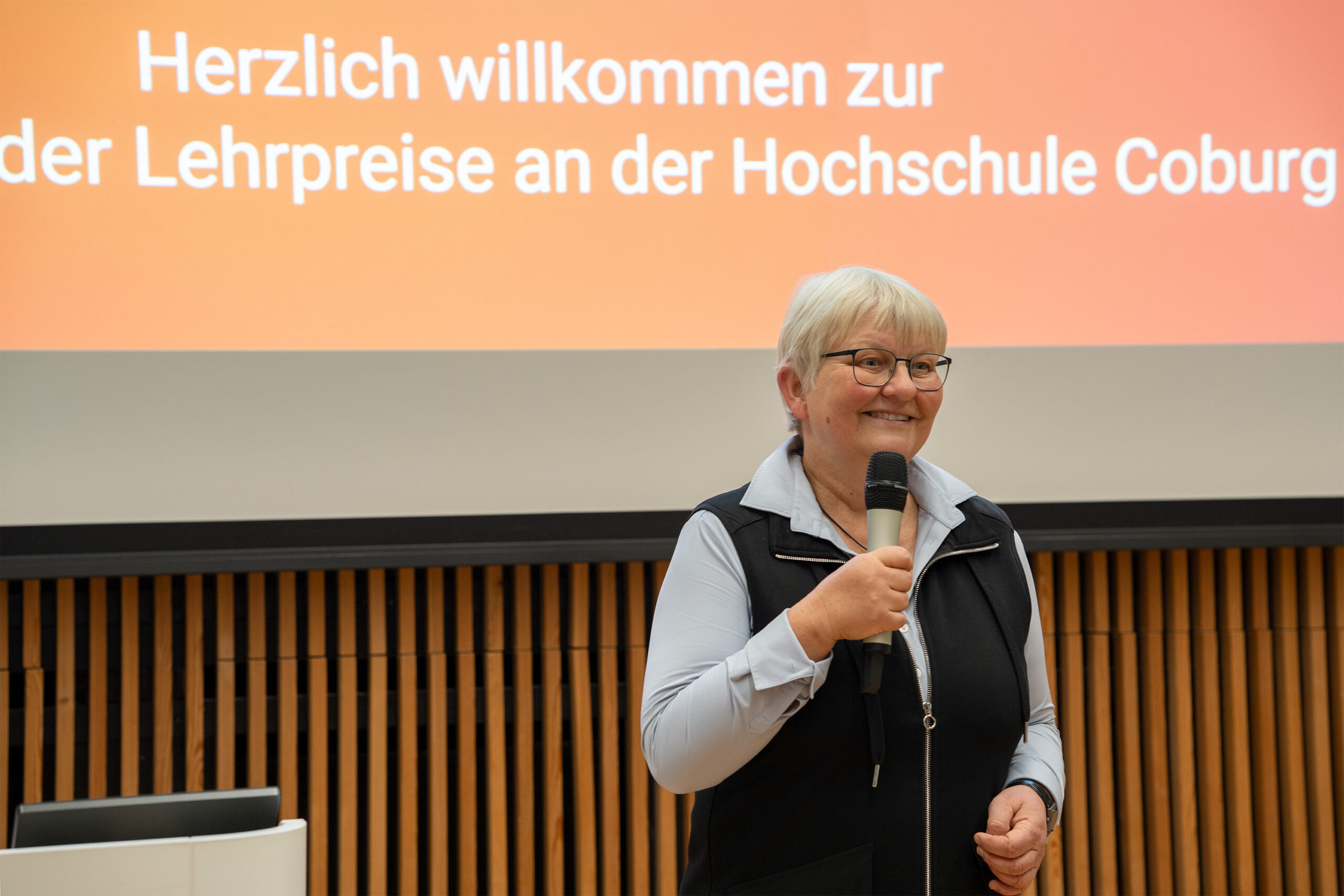 Eine ältere Frau mit kurzen grauen Haaren und Brille lächelt, während sie ein Mikrofon hält. Sie steht vor einem Bildschirm mit einem deutschen Text, der die Lehrenden zur Verleihung des Preises für innovative Lehre der Hochschule Coburg begrüßt. at Hochschule Coburg