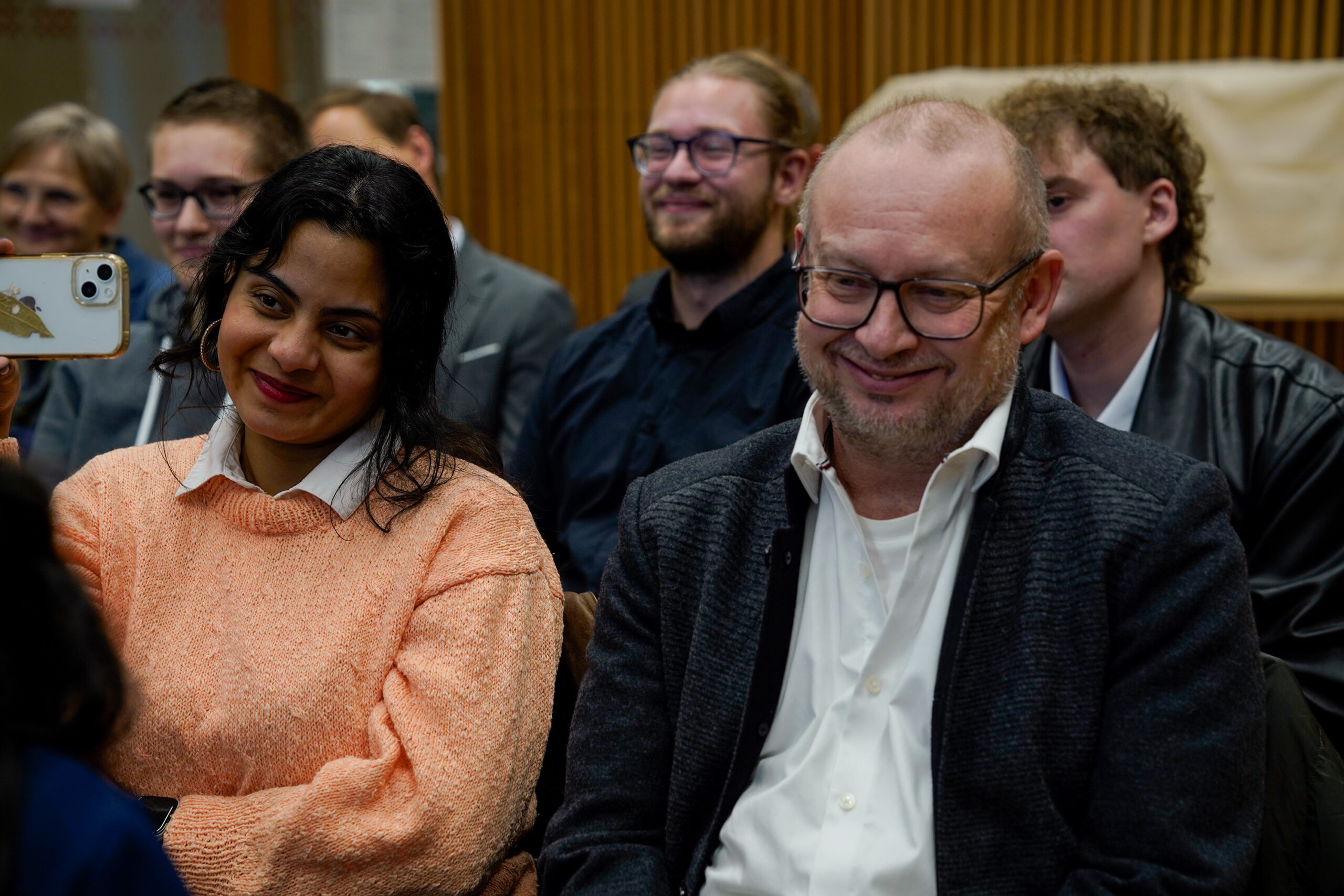 Eine Gruppe von Menschen sitzt eng beieinander, lächelt und beobachtet etwas aus dem Off. Eine Frau in einem pfirsichfarbenen Pullover, möglicherweise eine Lehrende, hält ein Telefon hoch, um eine Aufnahme zu machen, während ein Mann mit Brille und weißem Hemd neben ihr lächelt - er hält innovative Momente in der Lehre fest. at Hochschule Coburg