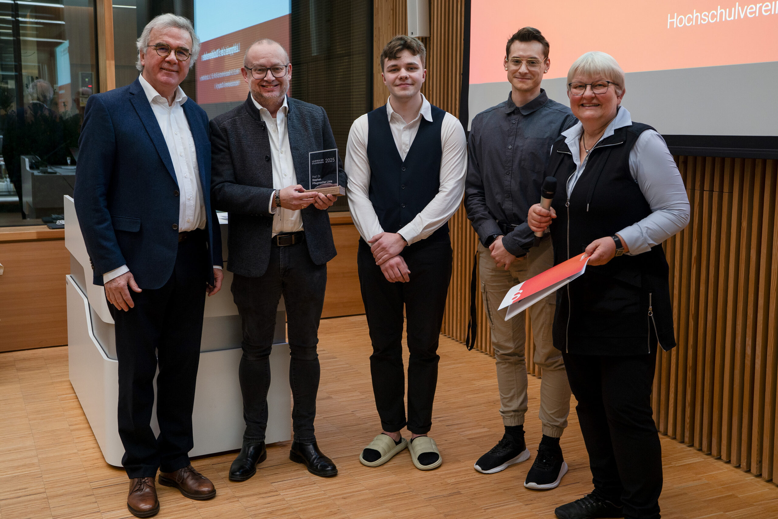 Fünf Personen stehen in einem Raum und posieren für ein Foto. Eine Person hält eine Auszeichnung für innovative Lehrende, eine andere hält ein Mikrofon und eine rote Mappe. Im Hintergrund sind eine Präsentationswand und eine Holzverkleidung zu sehen. at Hochschule Coburg