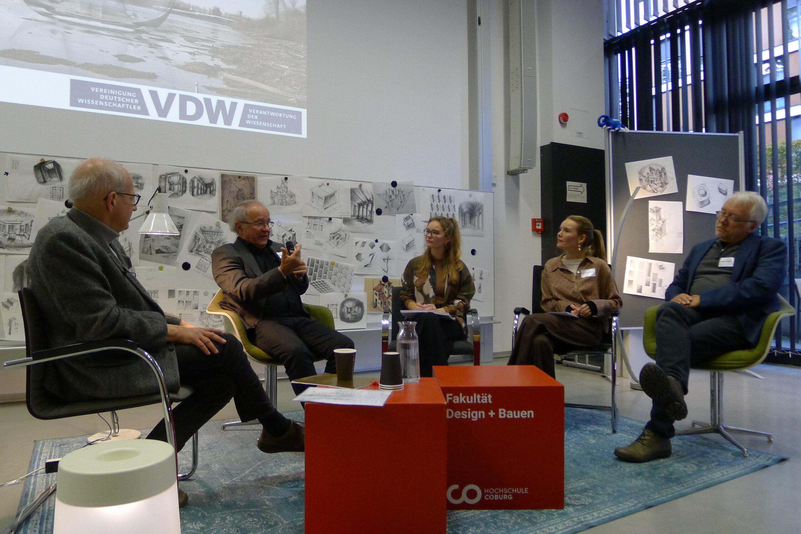 Fünf Personen sitzen im Halbkreis und diskutieren während des VDW-Aktionstages in der Hochschule auf einem Podium. Hinter ihnen hängen Zeichnungen und Skizzen, davor ein roter Würfel mit der Aufschrift "Fakultät Design + Bauen", der zum Räume fühlen einlädt. at Hochschule Coburg