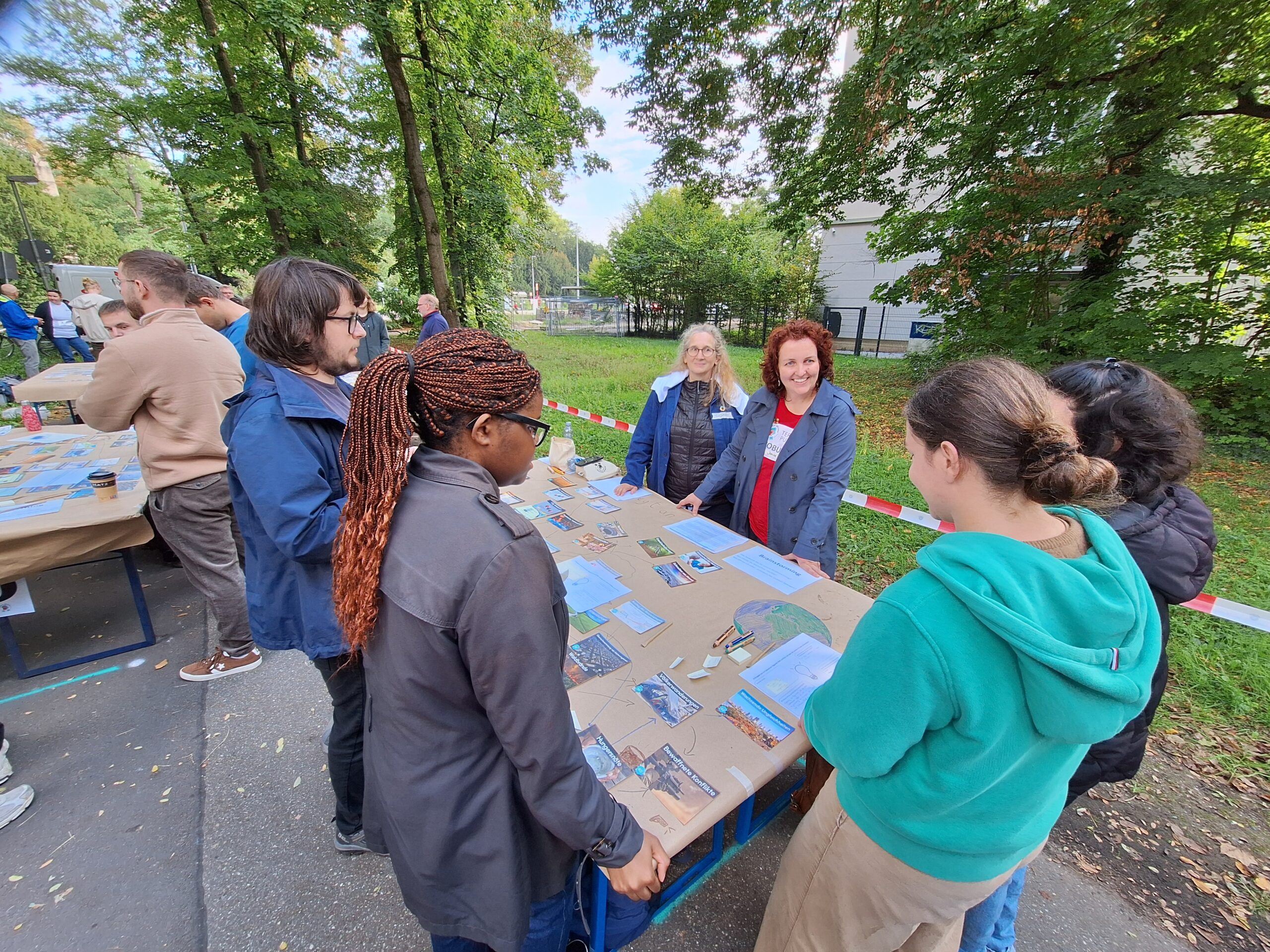 Eine Gruppe von Menschen steht um einen Tisch im Freien, der mit Postkarten und Fotos bedeckt ist, und unterhält sich über Klimabildung. Die Veranstaltung findet in einem parkähnlichen Gelände in der Nähe einer Hochschule statt, mit Bäumen und einem Gebäude im Hintergrund. at Hochschule Coburg
