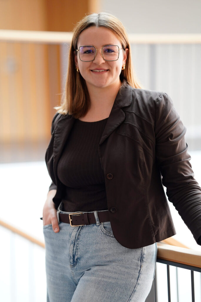 Eine Frau mit glattem, schulterlangem Haar, Brille und einem leichten Lächeln trägt einen dunklen Blazer, ein schwarzes Oberteil und hellblaue Jeans. Sie lehnt lässig an einem Geländer in einem hellen Innenraum. at Hochschule Coburg