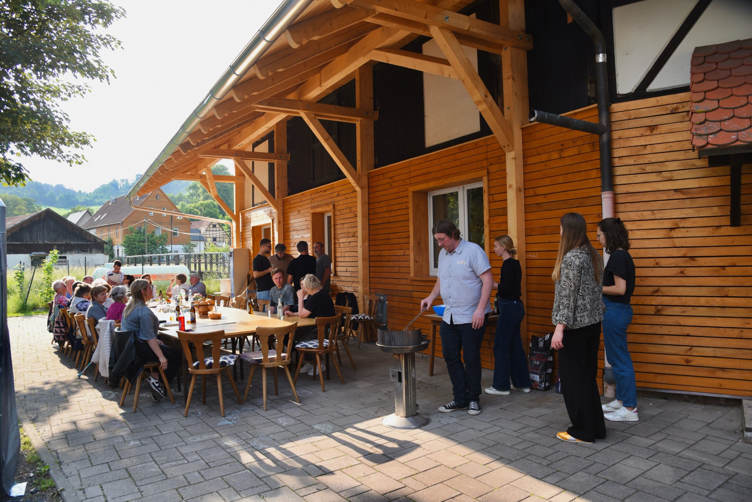 Eine Gruppe von Menschen versammelt sich um einen langen Tisch im Freien, der für eine Mahlzeit neben einem Holzgebäude gedeckt ist. Sie genießen das Essen und unterhalten sich unter einer hölzernen Markise. Automatisch gespeicherter Entwurf: Es ist ein sonniger Tag, an dem man sich angeregt unterhält. at Hochschule Coburg