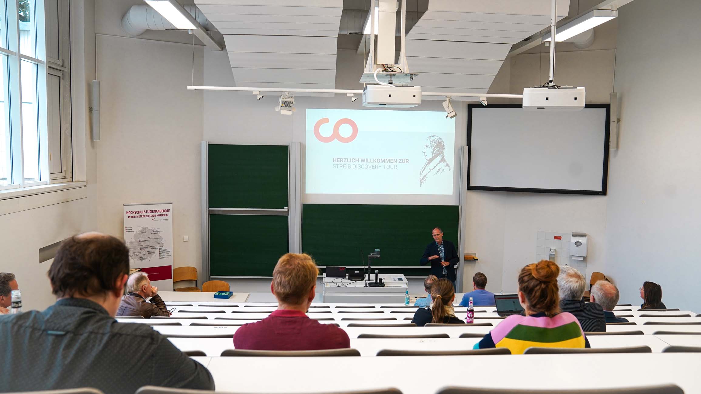 Ein Dozent steht vorne in einem Klassenzimmer der Hochschule Coburg und spricht zu den in Reihen sitzenden Studenten. Ein Bildschirm zeigt eine Begrüßungsbotschaft und eine Grafik, daneben Kreidetafeln und Geräte, die den Rahmen für Wissenschaft und Lernen bilden. at Hochschule Coburg