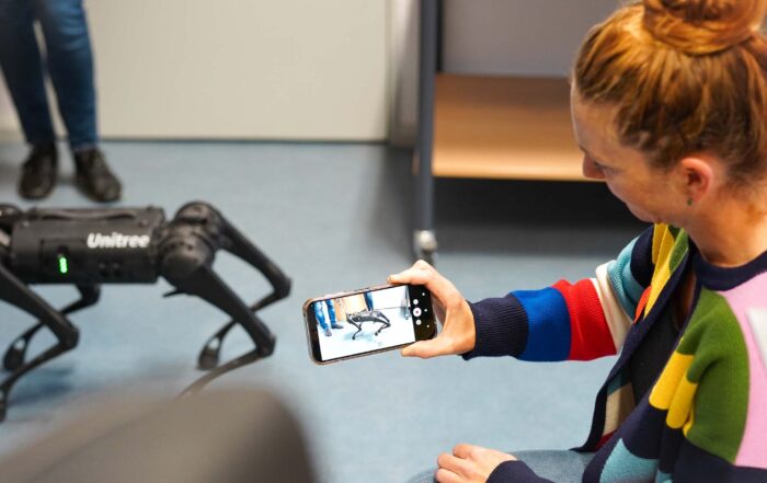 Eine Frau in einem farbenfrohen Pullover fotografiert einen schwarzen, vierbeinigen Unitree-Roboterhund mit ihrem Smartphone an der Hochschule Coburg und fängt damit die Schnittstelle zwischen Wissenschaft und Technik in einem Raum ein, während eine weitere Person im Hintergrund steht. at Hochschule Coburg