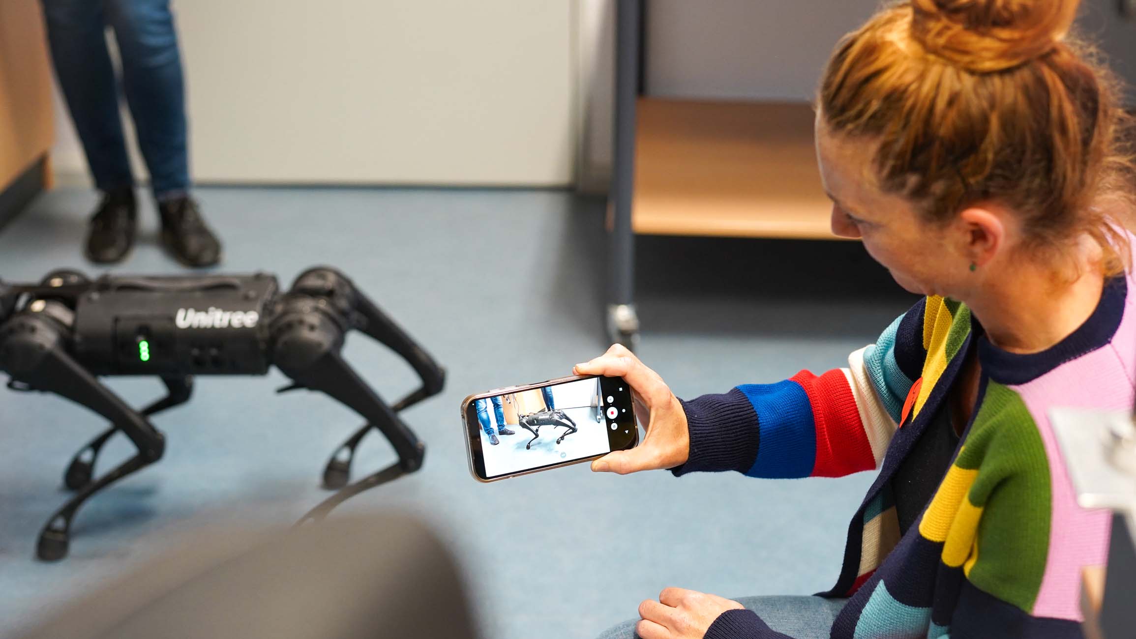 Eine Frau in einem farbenfrohen Pullover fotografiert einen schwarzen, vierbeinigen Unitree-Roboterhund mit ihrem Smartphone an der Hochschule Coburg und fängt damit die Schnittstelle zwischen Wissenschaft und Technik in einem Raum ein, während eine weitere Person im Hintergrund steht. at Hochschule Coburg