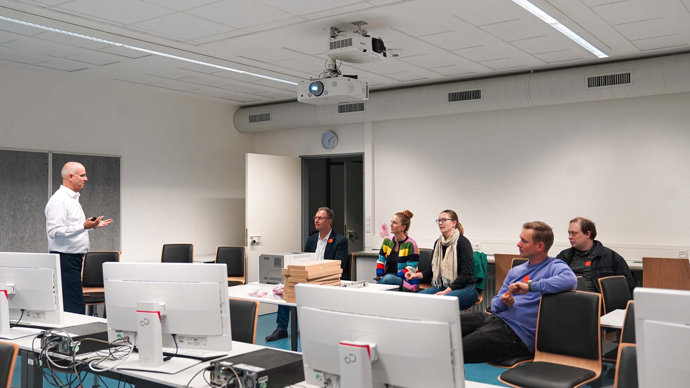 Ein Mann steht vorne in einem modernen, gut beleuchteten Klassenzimmer der Hochschule Coburg und spricht zu fünf sitzenden Erwachsenen. Auf den Tischen im Vordergrund stehen mehrere Desktop-Computer, und von der Decke hängt ein Projektor. at Hochschule Coburg