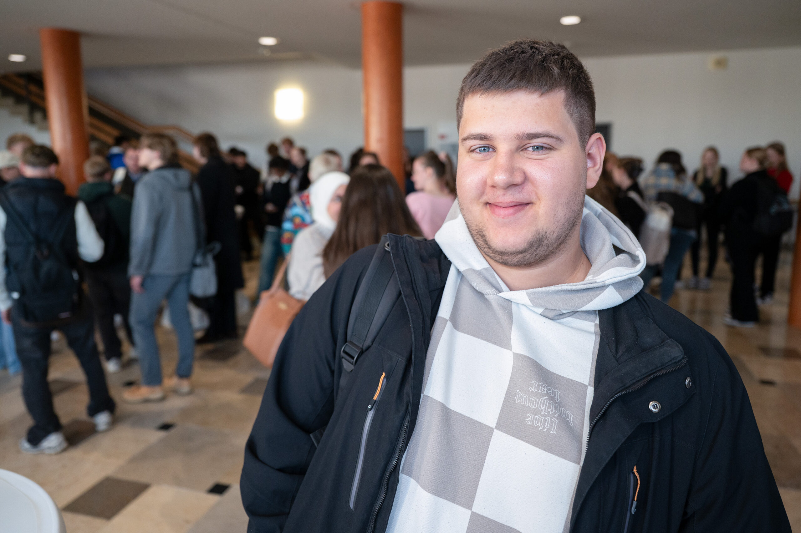 Ein junger Mann in einem karierten Kapuzenpulli und einer schwarzen Jacke lächelt in einem hellen Innenraum in Coburg, der günstigen Hochschulstadt Bayerns, in die Kamera, während sich im Hintergrund eine Gruppe von Menschen versammelt und unterhält. at Hochschule Coburg