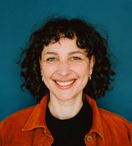 A woman with curly dark hair and blue eyes smiles warmly at the camera. She is wearing a rust-colored jacket over a black top, embodying confidence in her choice to study while working. The background is a solid teal blue. at Hochschule Coburg