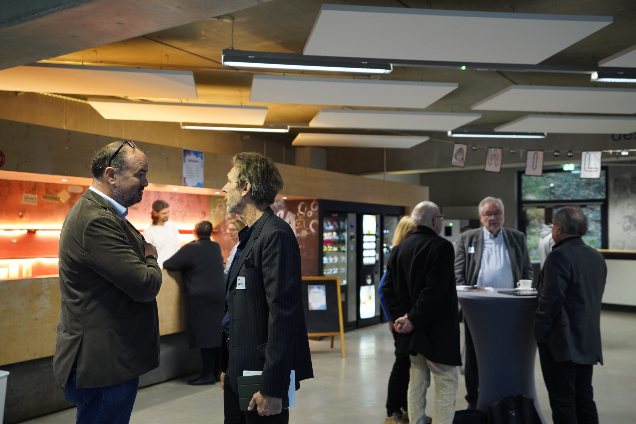 Mehrere Männer in Business-Kleidung stehen während des VDW-Aktionstages in einem modernen, offenen Innenraum der Hochschule und unterhalten sich. Einige haben sich um einen hohen Tisch in der Nähe des Verkaufsbereichs und der Café-Theke versammelt und schaffen so eine professionelle und doch lockere "Räume fühlen"-Atmosphäre. at Hochschule Coburg