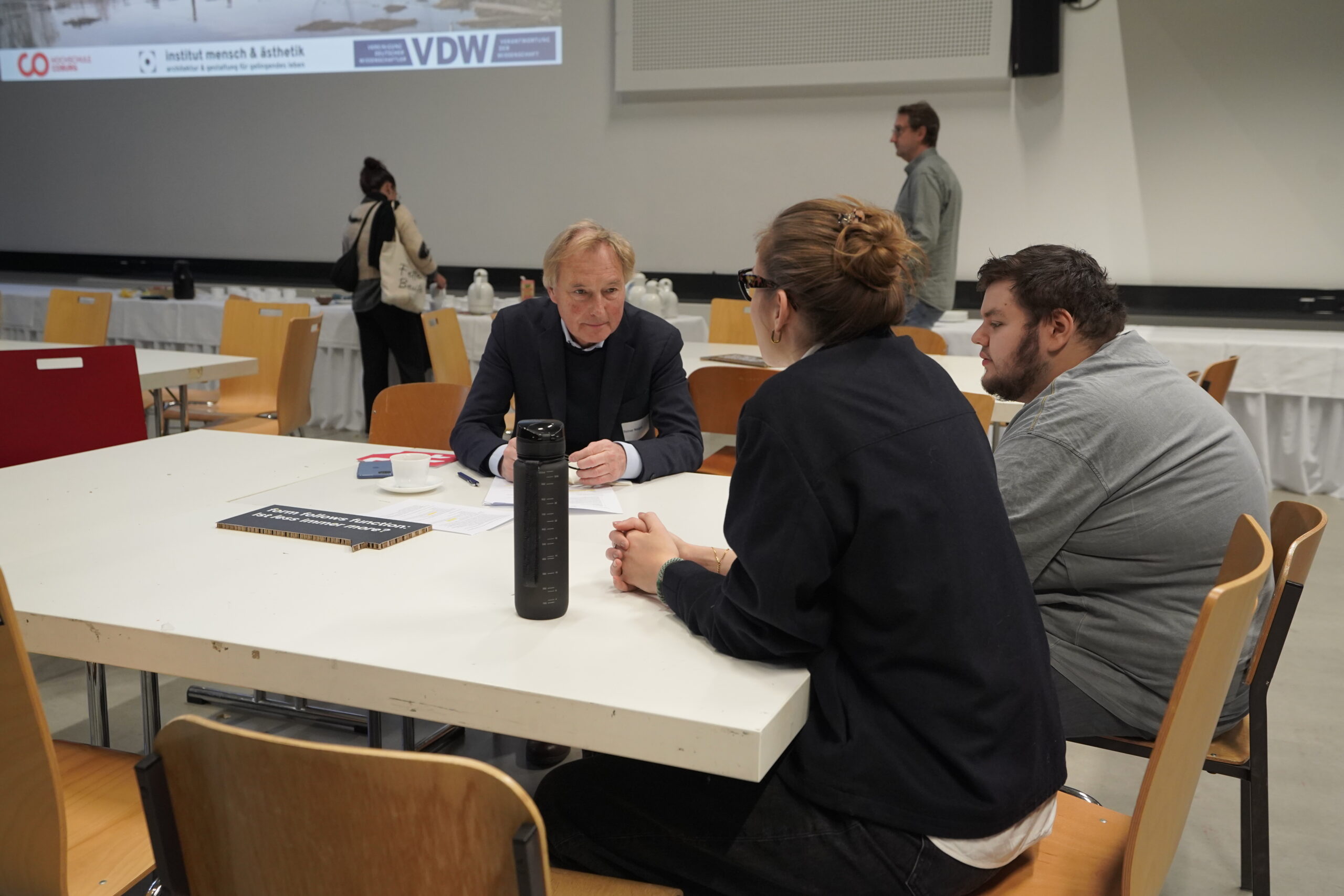 Drei Personen sitzen während des VDW-Aktionstages in einem Konferenzraum der Hochschule an einem Tisch und diskutieren, im Hintergrund stehen weitere Personen und leere Stühle. An der Wand werden auf einem großen Bildschirm Präsentationsfolien gezeigt. at Hochschule Coburg