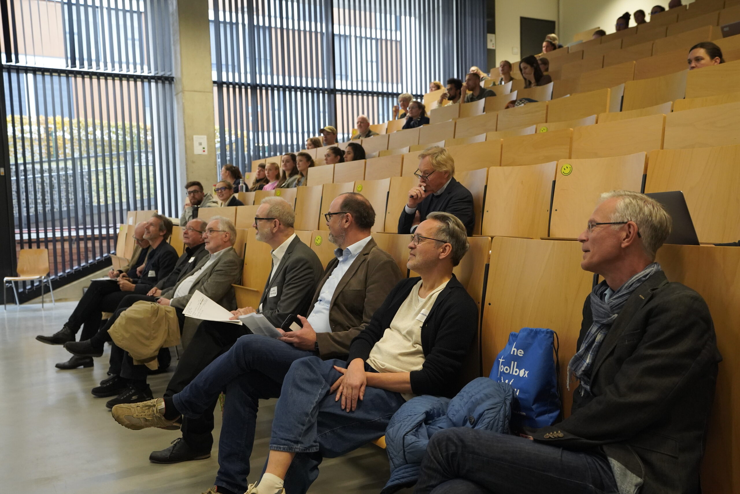 Die Menschen sitzen verstreut in einem Hörsaal der Hochschule mit Holzstühlen und hören aufmerksam zu. Die meisten Zuhörer sind ältere Erwachsene, einige halten Papiere oder Taschen in der Hand, und durch große Fenster am Raum strömt natürliches Licht herein. at Hochschule Coburg