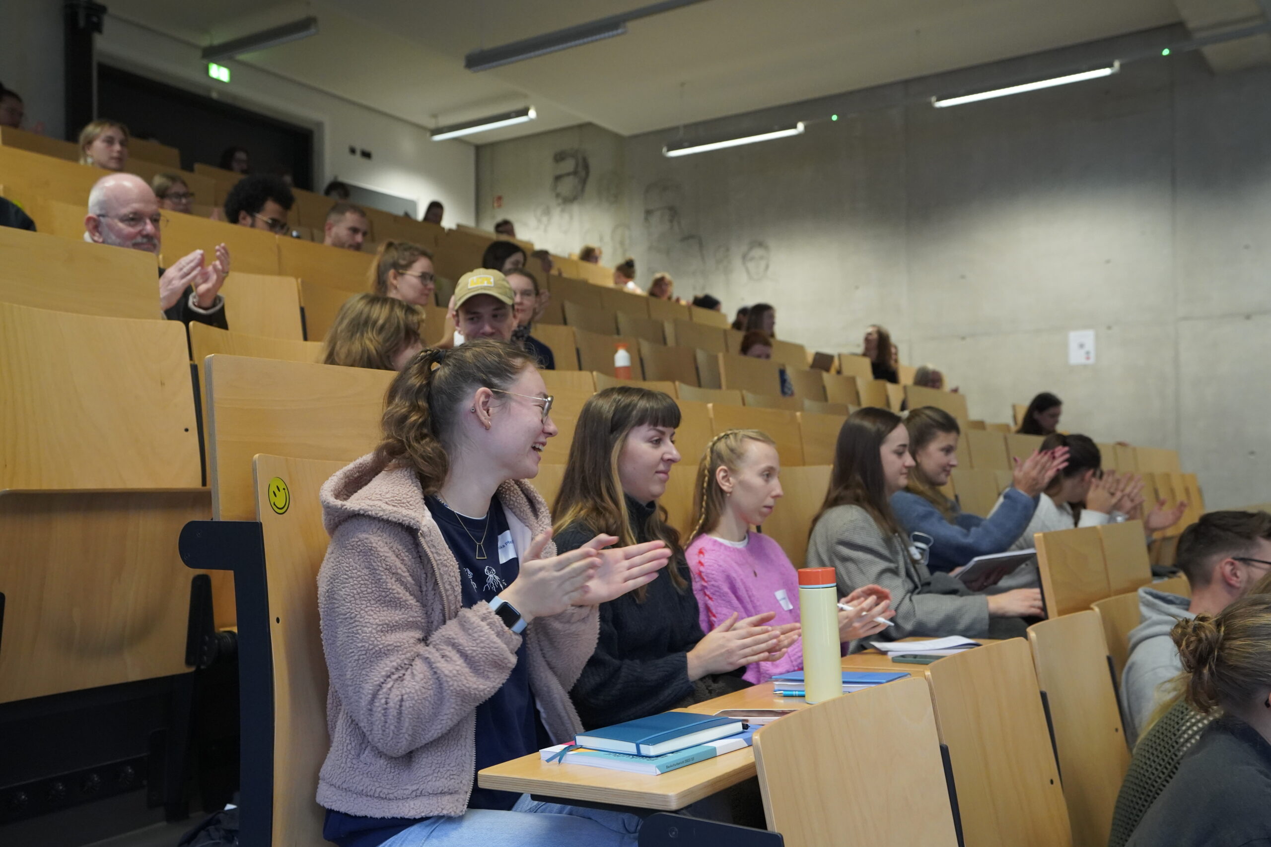 Studenten sitzen in einem Hörsaal der Hochschule mit Holzstühlen, klatschen und passen auf. Auf einigen Tischen liegen Notebooks und Wasserflaschen. Die Räume haben Betonwände und sind während des VDW-Aktionstages mit natürlichem Licht gefüllt. at Hochschule Coburg