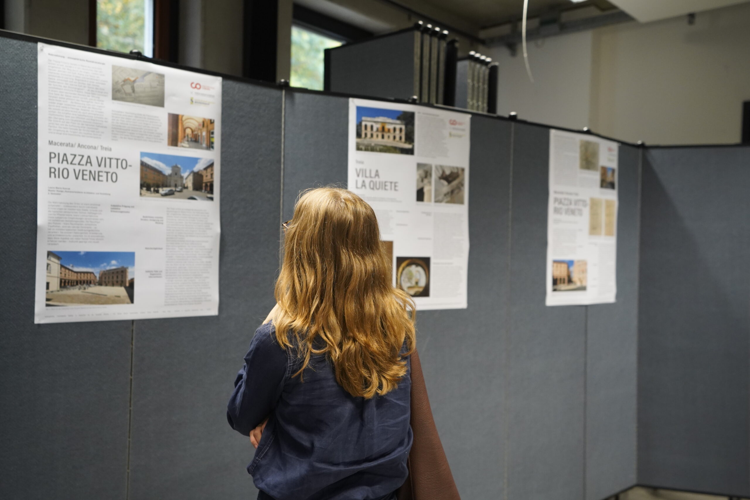 Eine Person mit langen, hellbraunen Haaren liest auf dem VDW-Aktionstag in einer gut beleuchteten Halle der Hochschule Informationsplakate über Architektur oder Gebäude und erkundet innovative Räume. at Hochschule Coburg