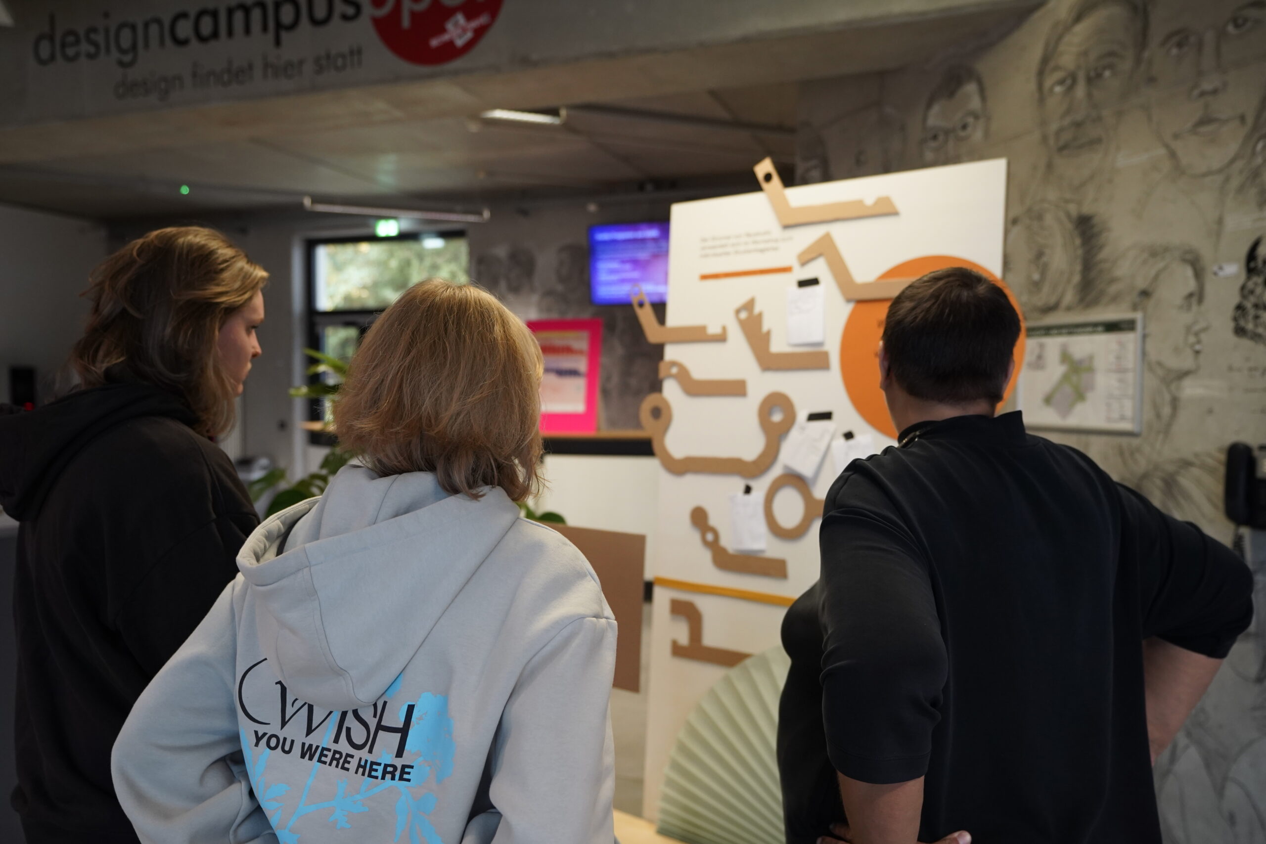 Drei Personen stehen in einem Raum und betrachten aufmerksam eine große Tafel mit Diagrammen und Papieren. Die kreativen Räume suggerieren ein Bildungsumfeld, mit Skizzen an der Wand und "designcampus"-Schildern, die auf eine Hochschulveranstaltung oder einen VDW-Aktionstag hindeuten. at Hochschule Coburg