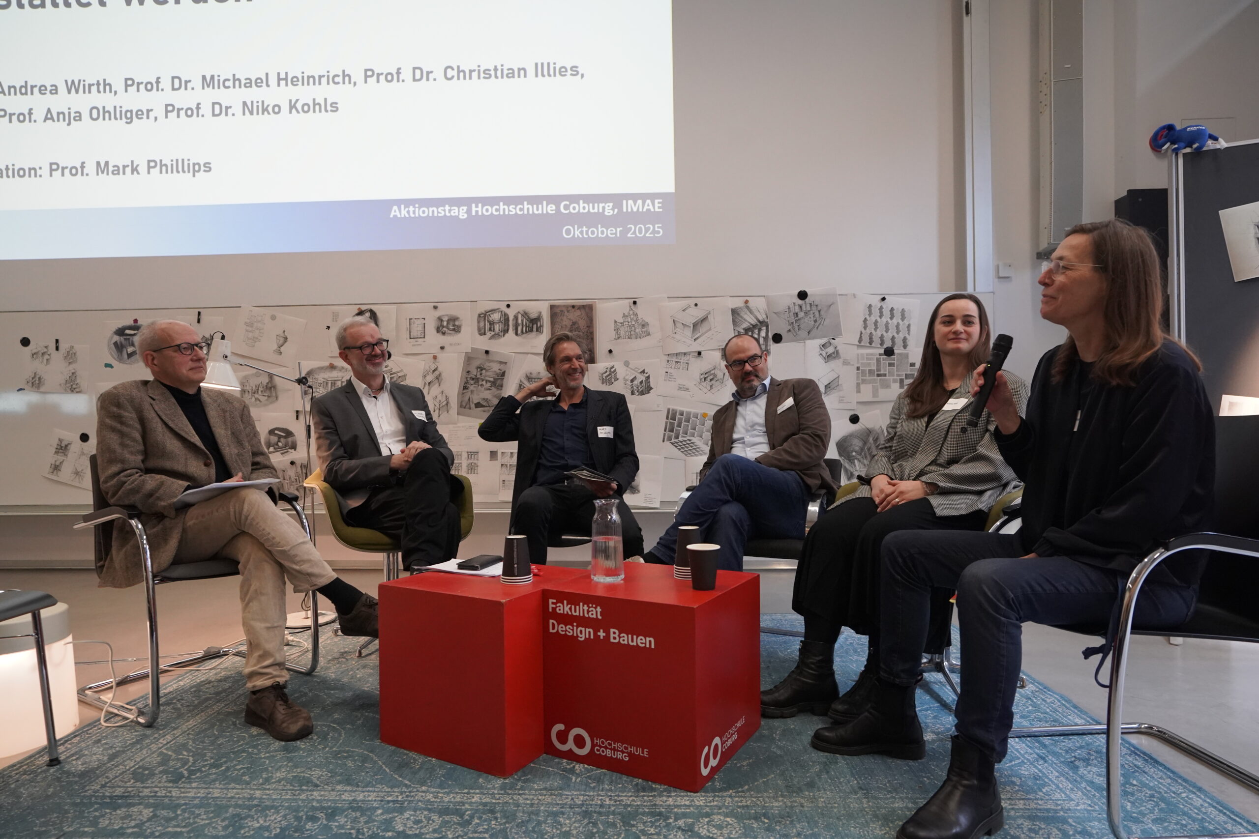 Sechs Personen sitzen bei einer Podiumsdiskussion in einer Hochschule und unterhalten sich. In dem modernen Raum hängen Skizzen an den Wänden, auf dem Tisch liegen Namenskarten, dahinter wird eine Präsentation des VDW-Aktionstags projiziert. at Hochschule Coburg