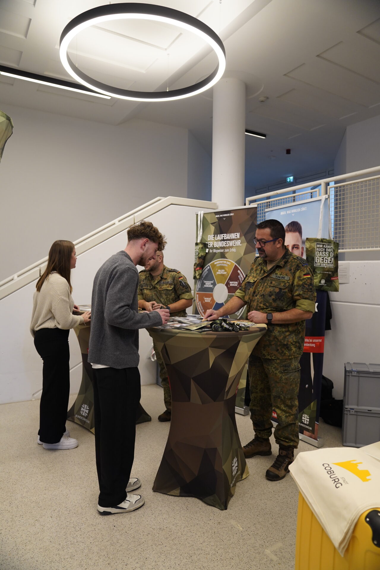 Zwei Männer in Militäruniformen stehen an einem Infostand und unterhalten sich mit zwei jungen Erwachsenen. Der militärische Stand mit einem bunten Spinnrad bietet Orientierung für die Zukunft auf der Coburger Studienmesse, möglicherweise während der Studienmesse 2025. at Hochschule Coburg