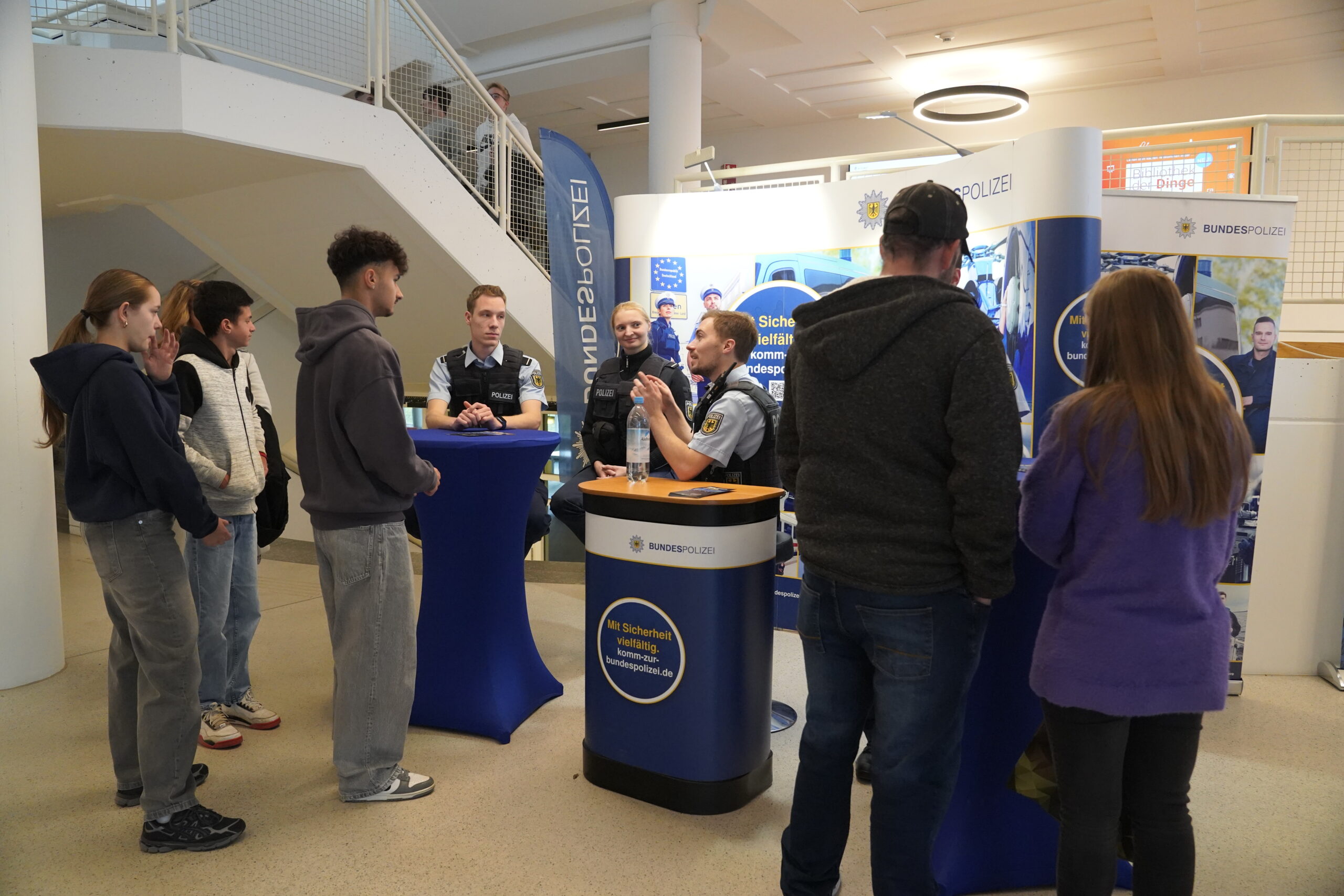 Mehrere Personen tauschen sich am Informationsstand der Bundespolizei während der Coburger Studienmesse aus. In einem hell erleuchteten Raum mit einer Treppe im Hintergrund informieren Beamte die Besucher über die Zukunft Orientierung neben Displays und Bannern. at Hochschule Coburg